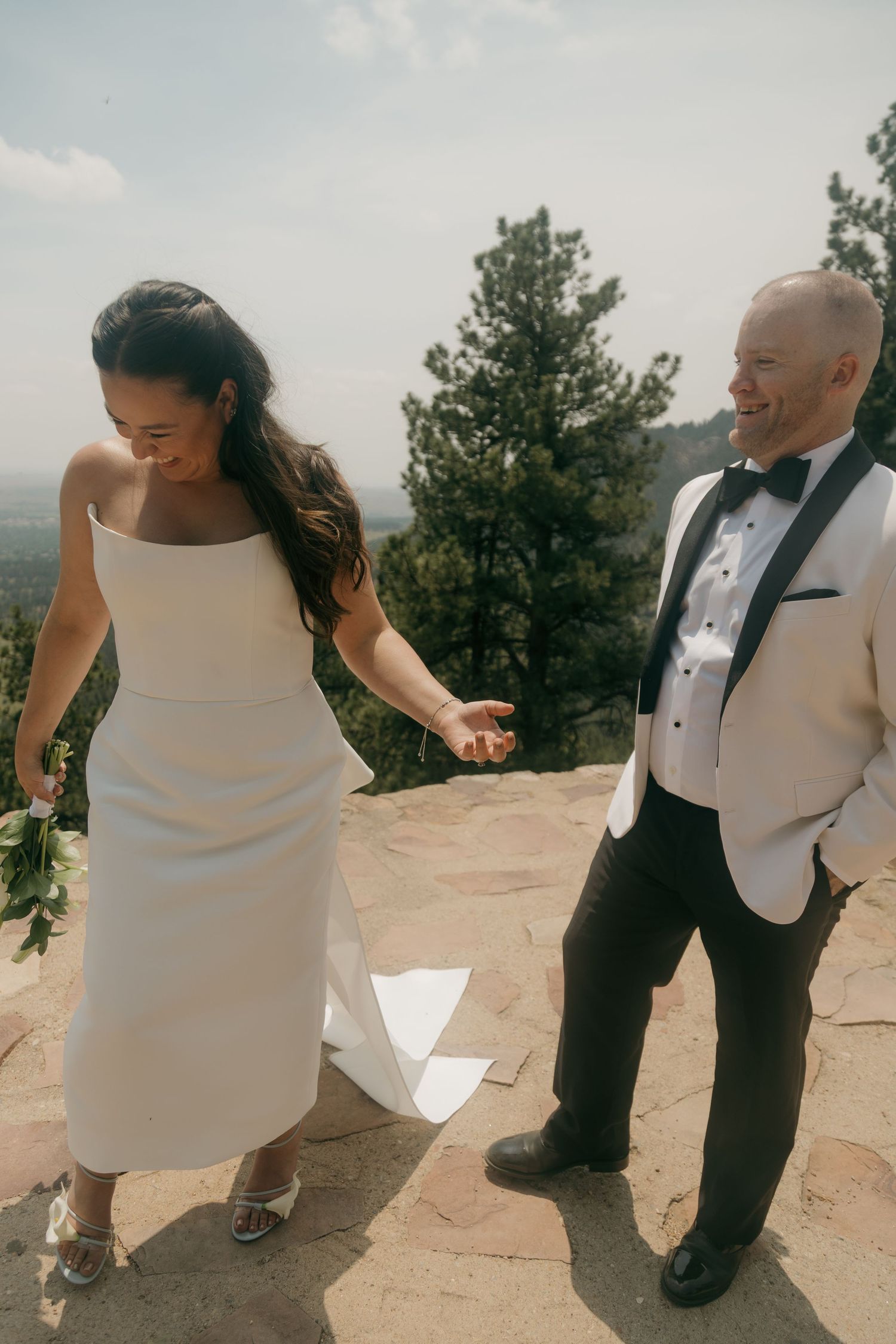 Bride and groom in elegant wedding attire hold hands and laugh on a rocky mountain overlook.