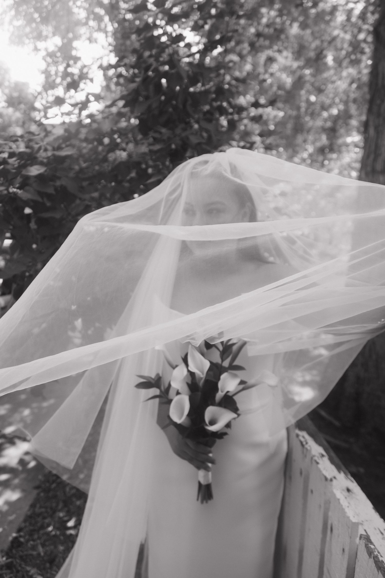Bride in white dress holding calla lily bouquet with veil blowing over face outdoors.