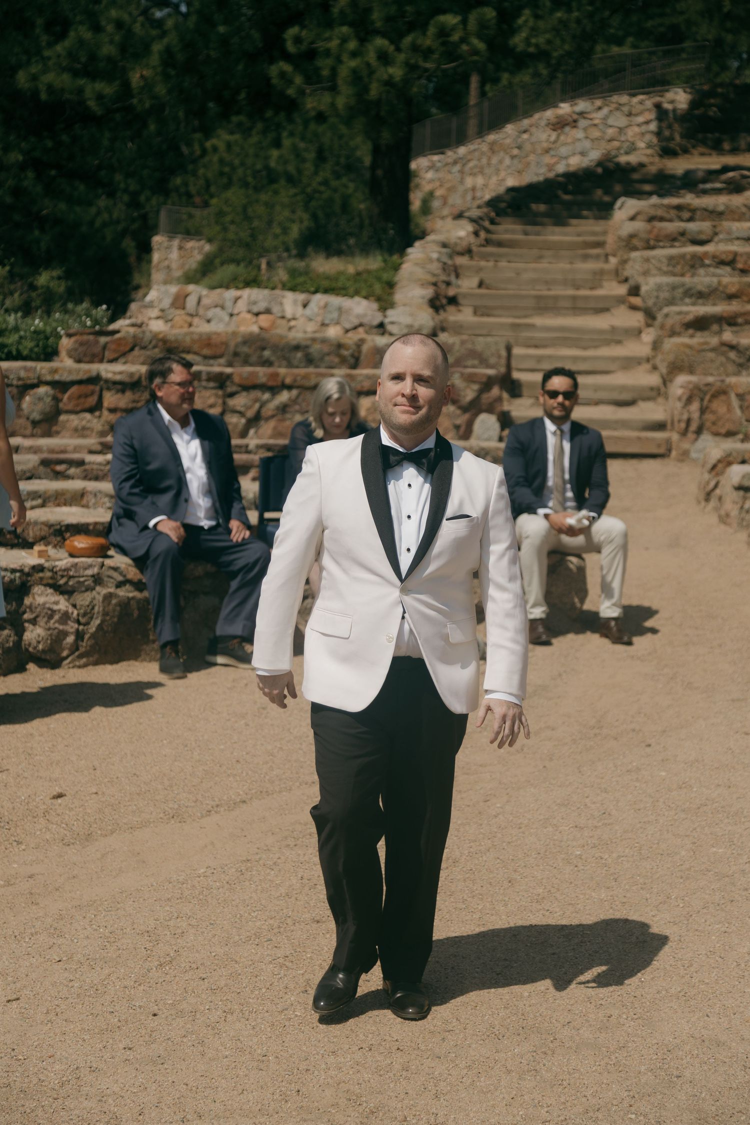 Groom in white tuxedo jacket walking confidently at an outdoor wedding venue with stone steps.