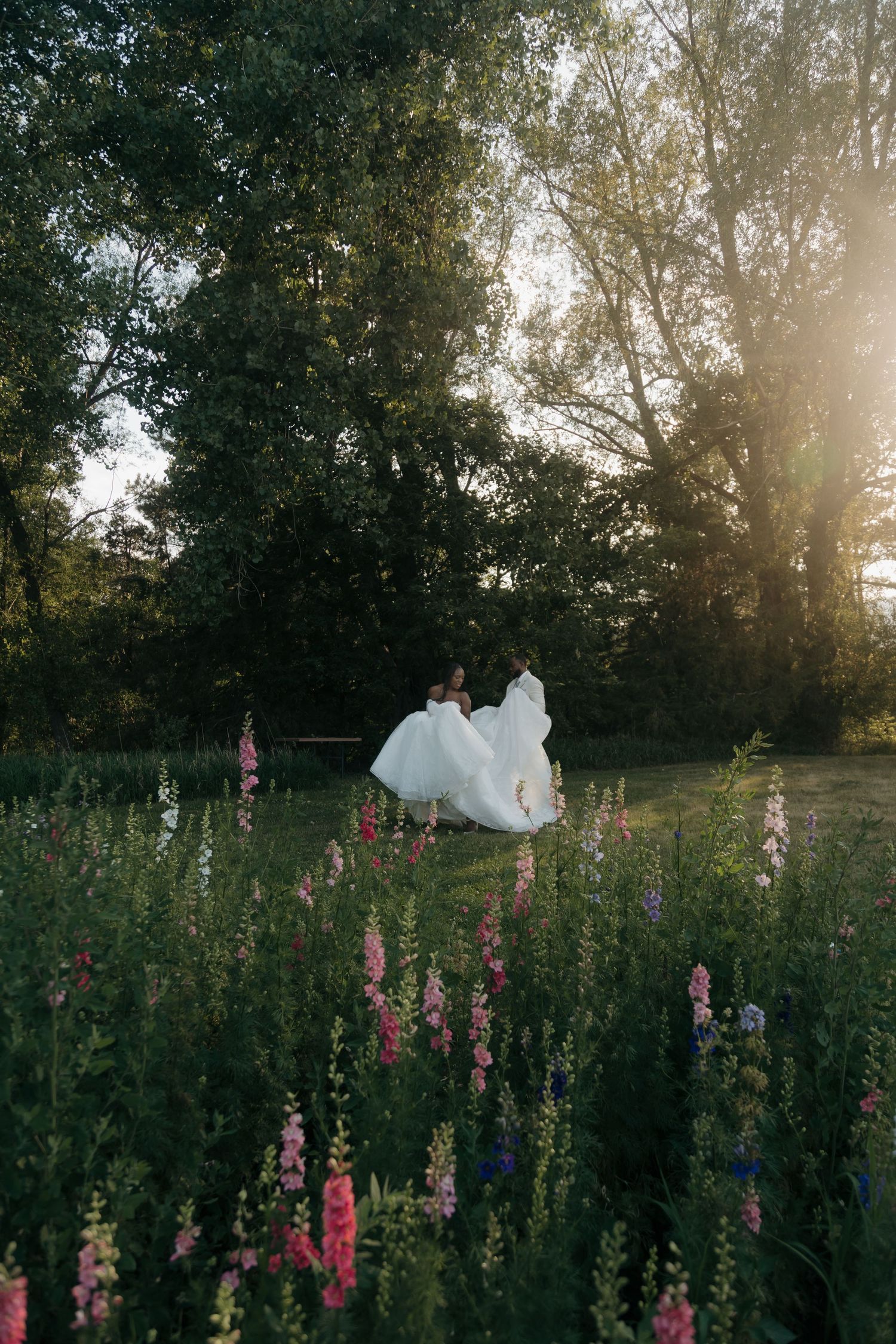 Bride in flowing white gown dancing in a wildflower meadow at golden hour with lush green trees.