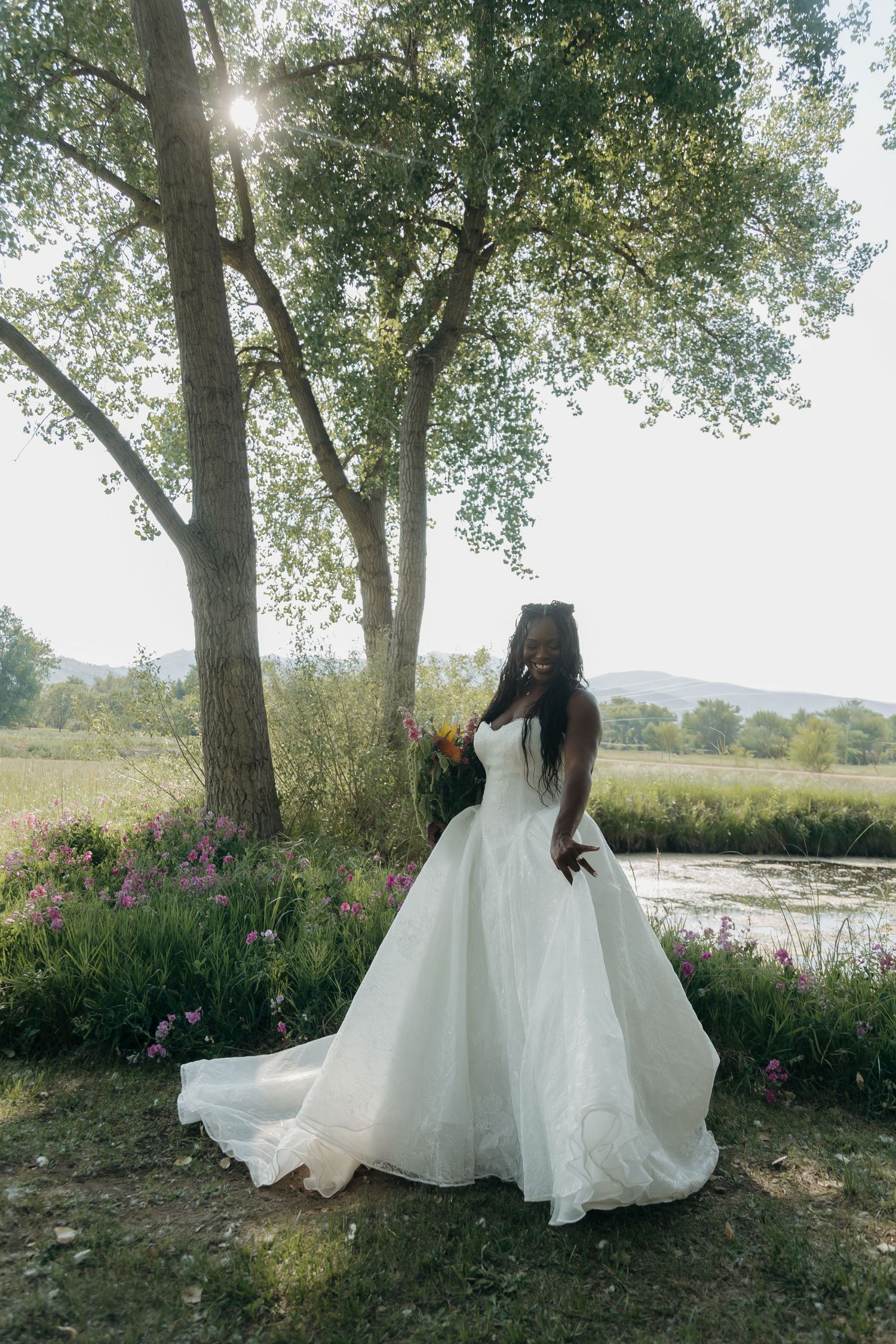 A Black bride in a flowing white ball gown holds a bouquet near a sunlit tree by a pond surrounded by wildflowers.