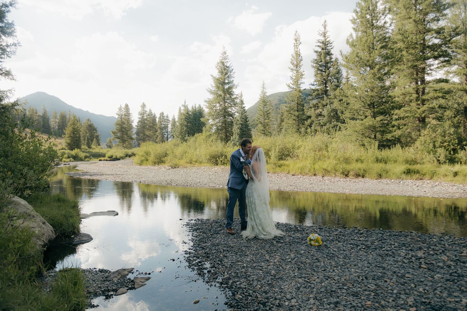 Bride and groom embrace on rocky riverbank surrounded by pine trees and mountains in a scenic outdoor wedding photo.