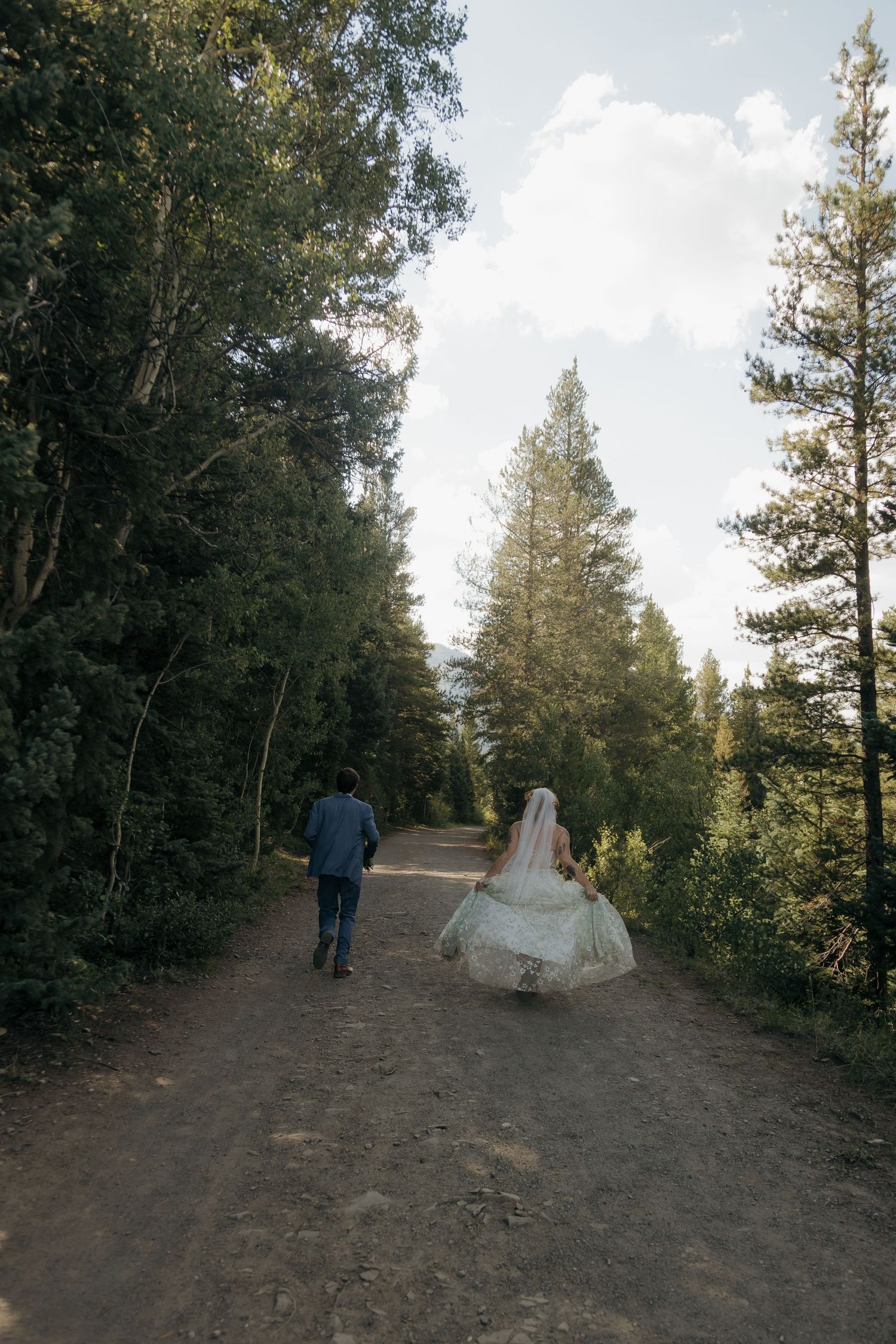 Bride and groom walking hand-in-hand down a forest dirt path surrounded by tall pine trees on their wedding day.
