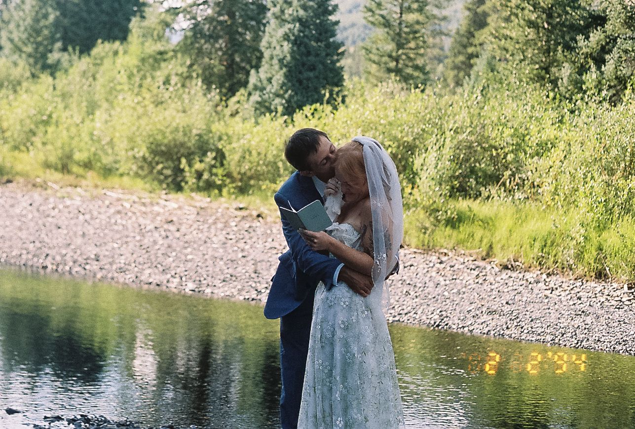 Bride and groom share first kiss by rocky riverbank surrounded by lush green summer foliage.