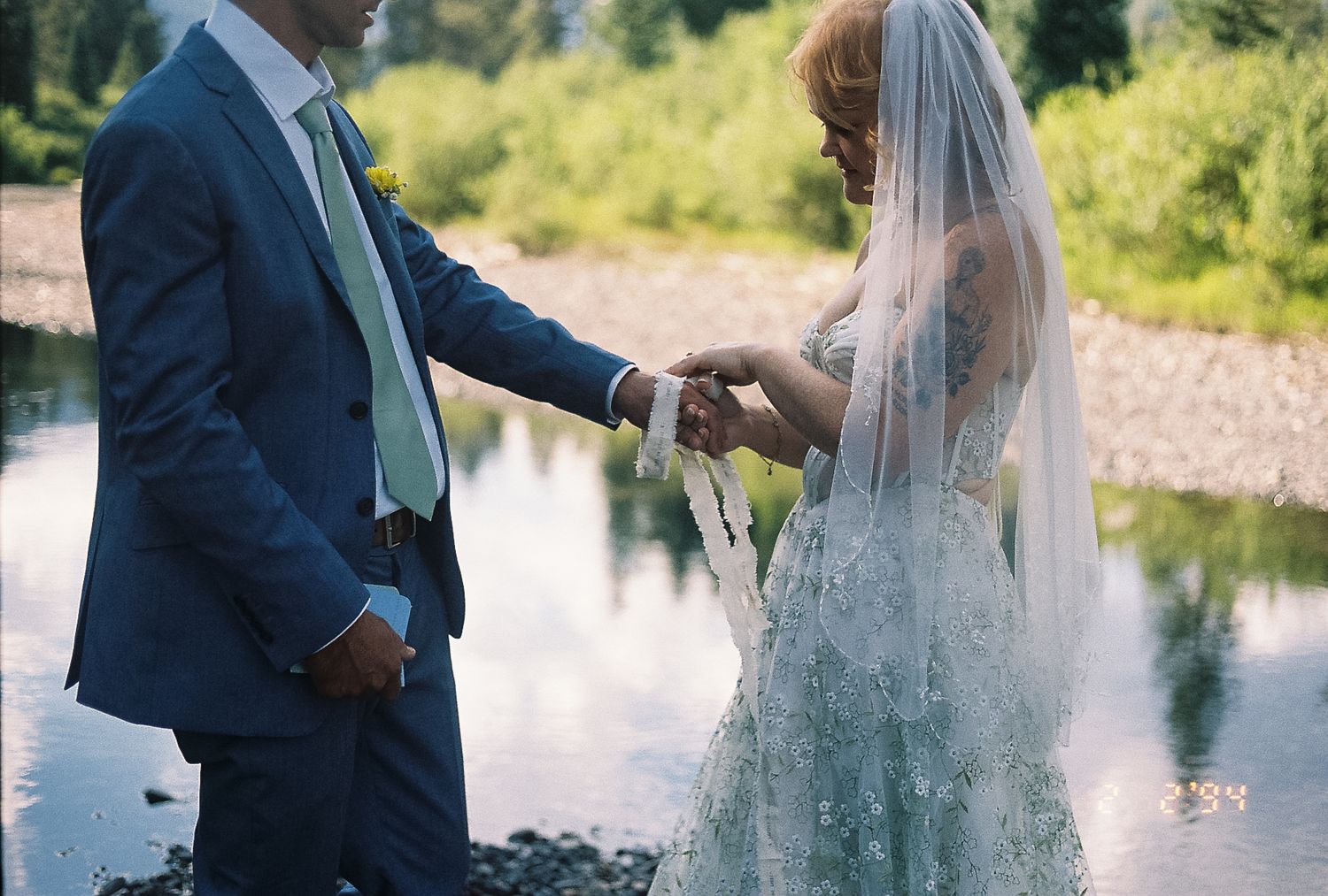 Bride in lace gown with veil and groom in blue suit exchanging vows by a riverside.