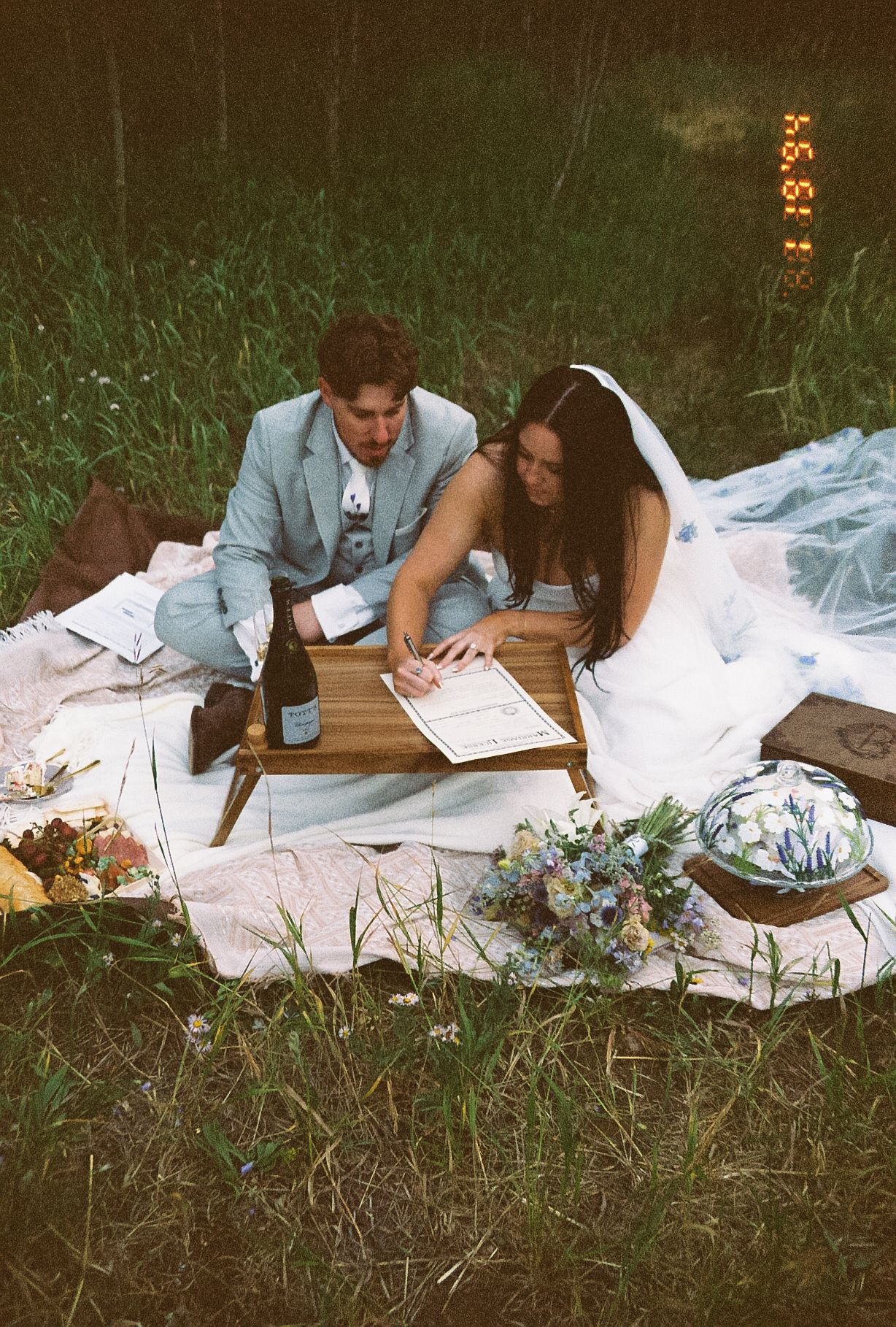 Bride and groom sign wedding certificate together on a picnic blanket in a meadow setting.