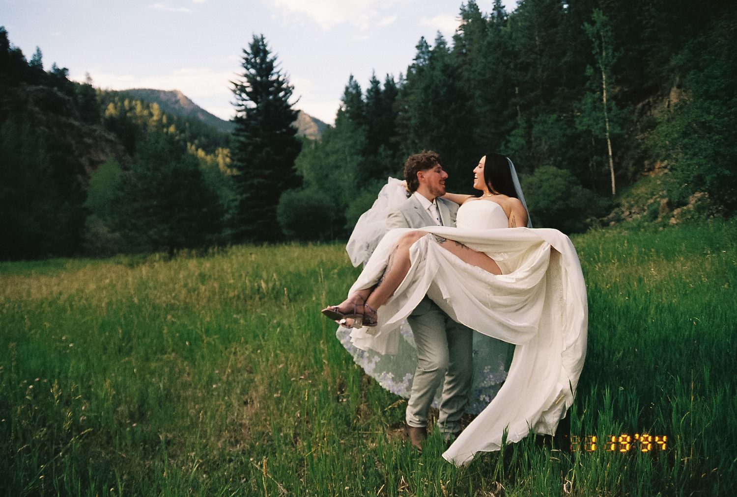 Groom carries laughing bride through a lush green mountain meadow in a film-style photo with vintage date stamp.