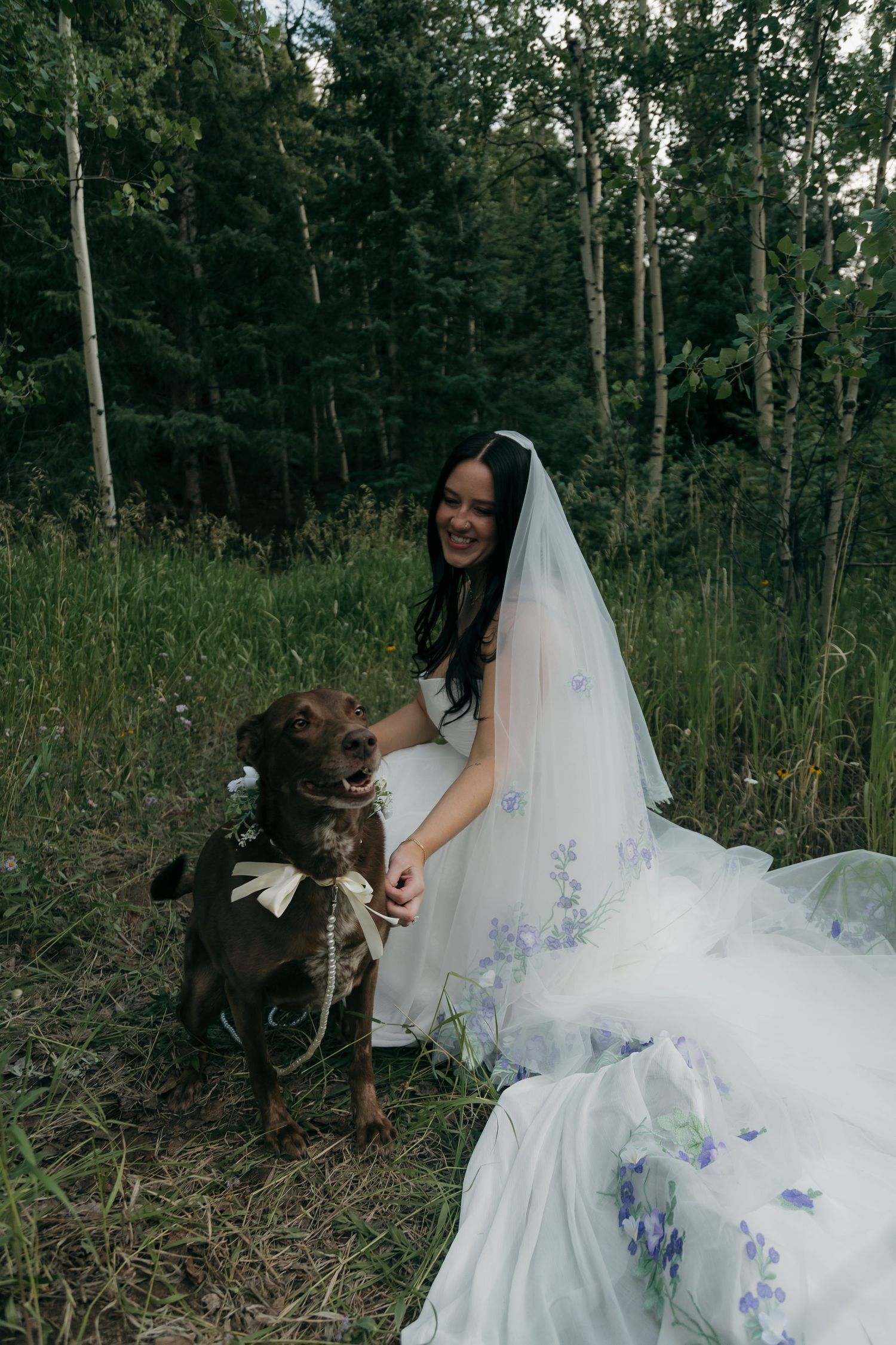 Smiling bride in white floral gown kneels beside her brown dog wearing a bow tie in a forest.