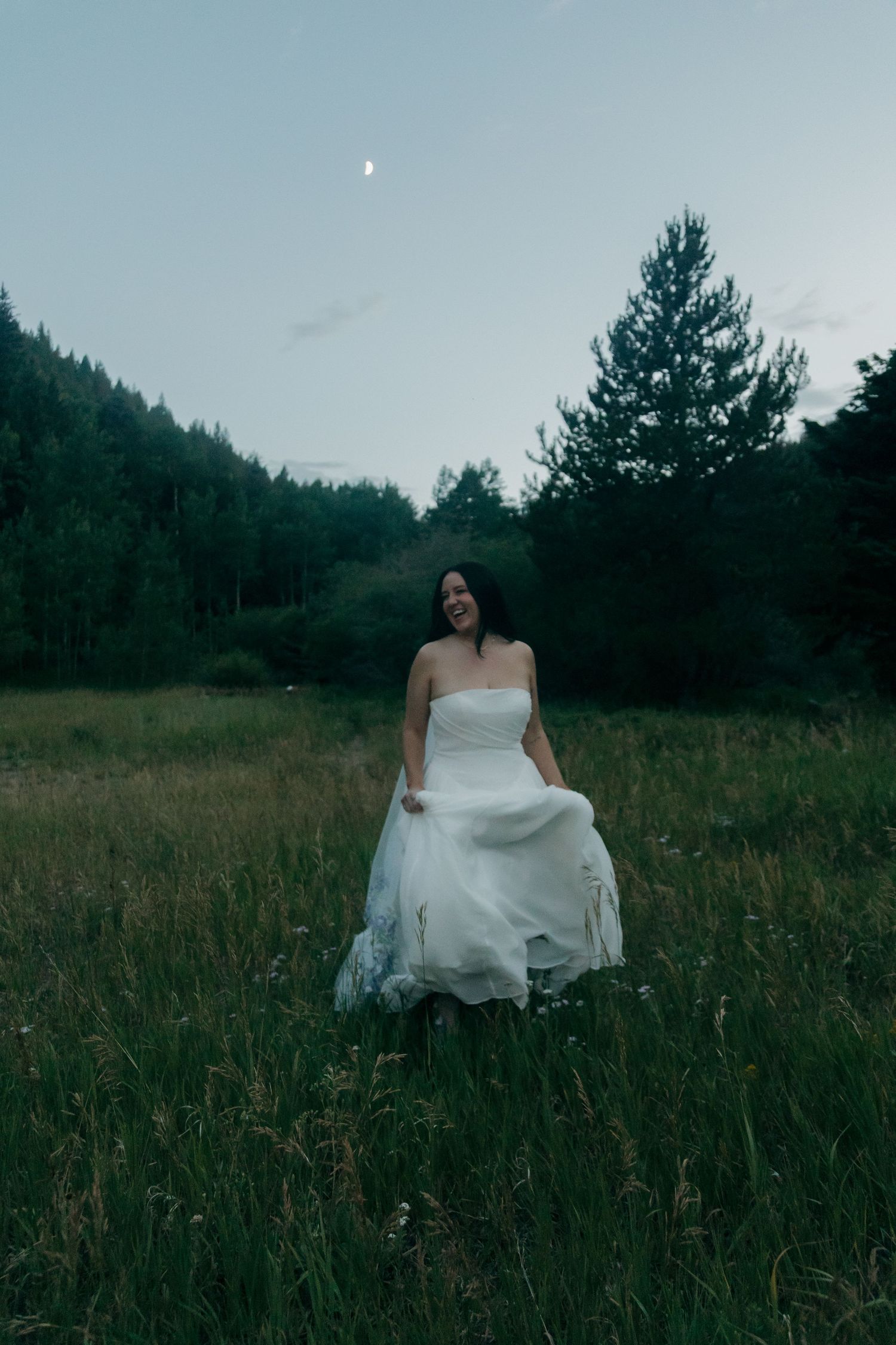 Smiling bride in strapless white gown walks through meadow under crescent moon at dusk.