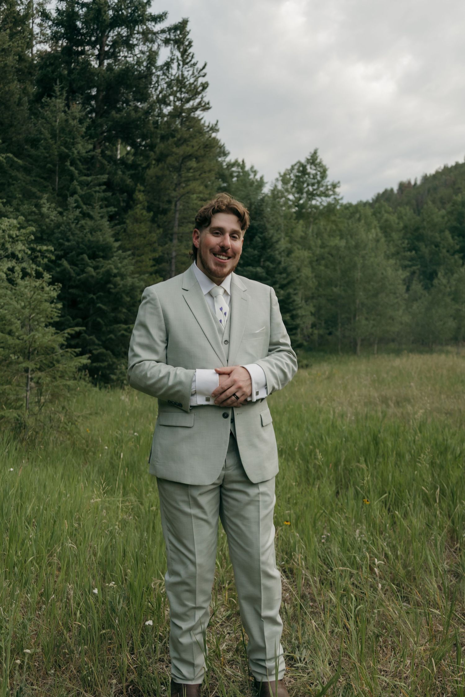 Smiling groom in light sage suit standing in a lush green meadow surrounded by forest trees.
