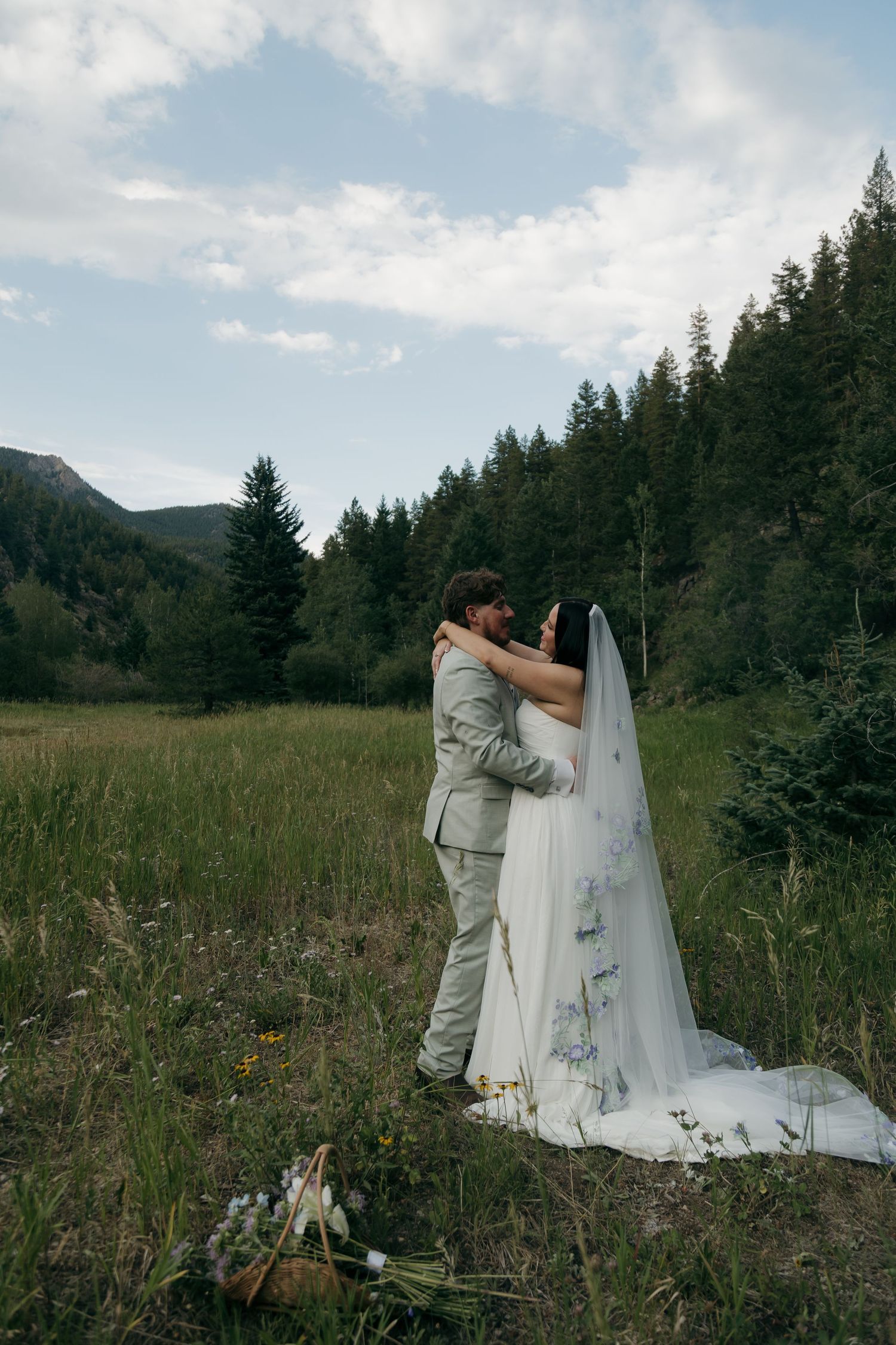 Wedding couple embraces in a scenic mountain meadow surrounded by tall evergreen trees and wildflowers.