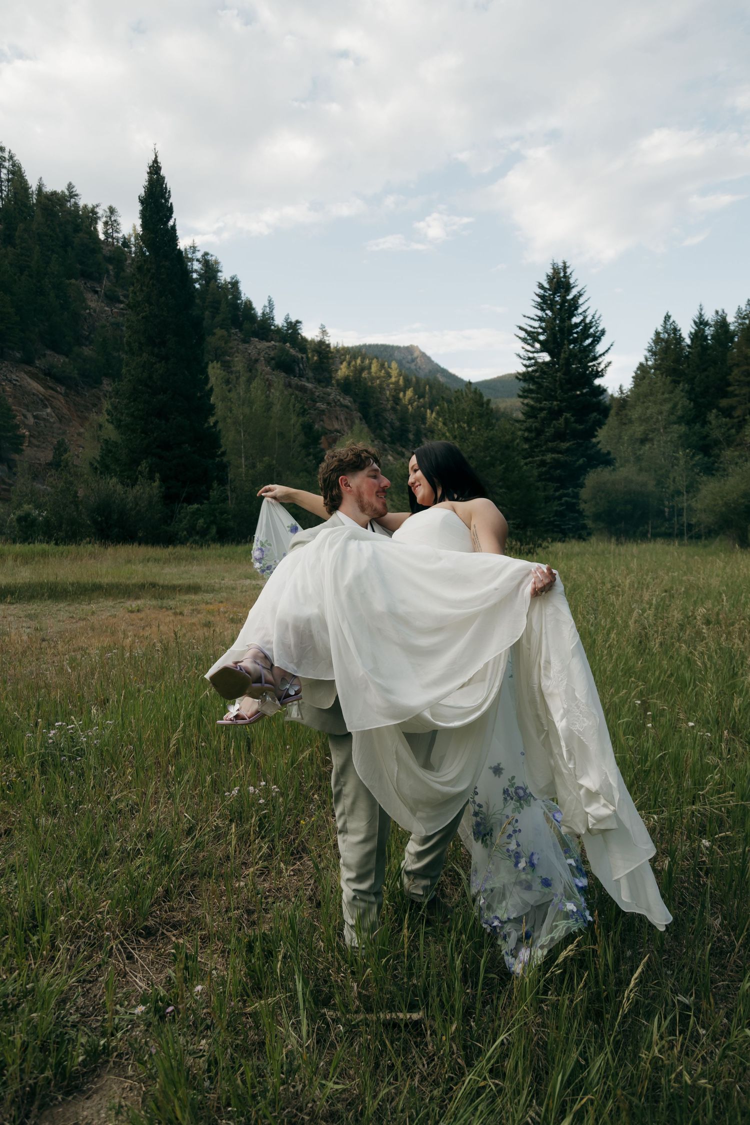 Groom in gray suit carries bride in white floral wedding dress through a green mountain meadow.