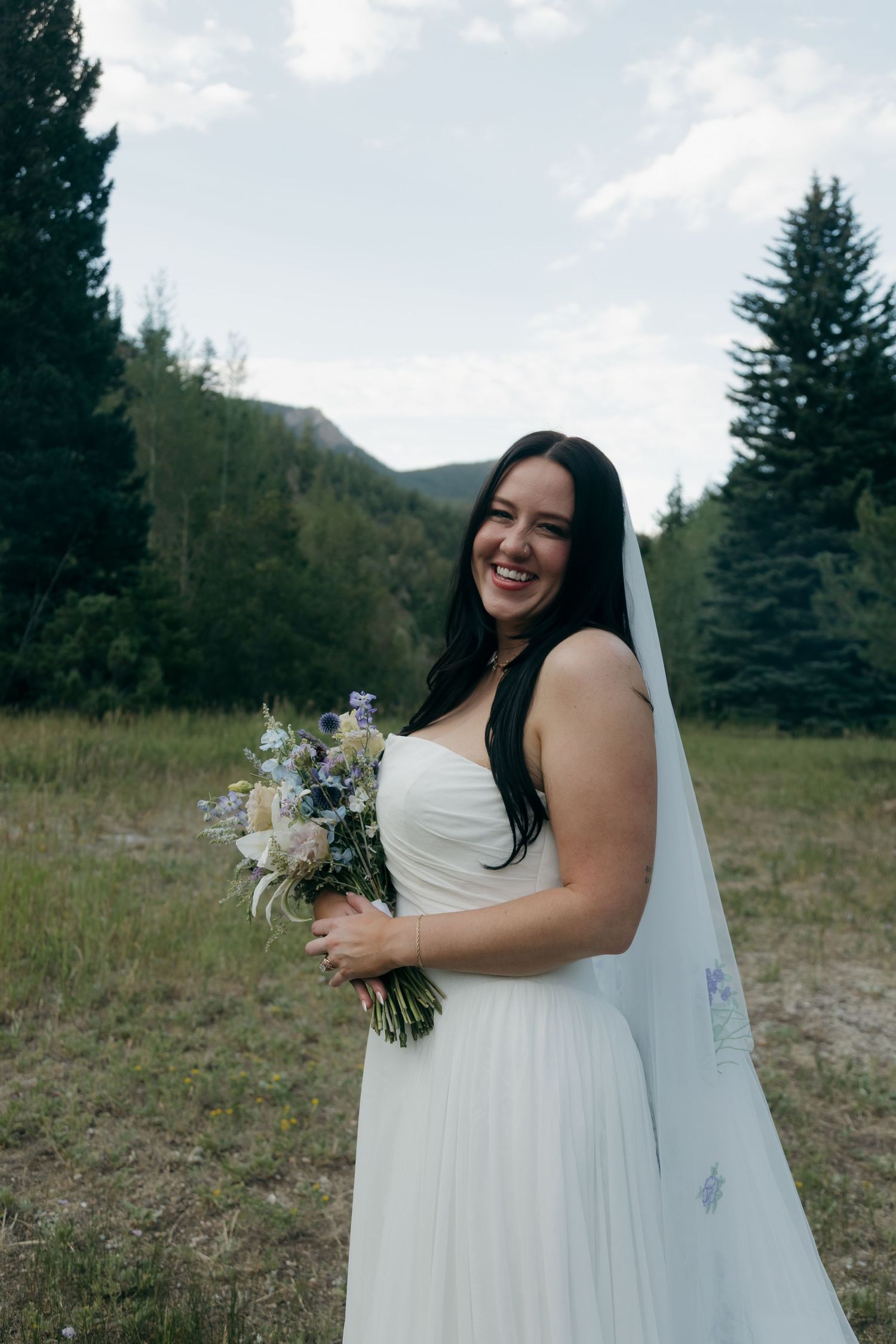 Smiling bride in strapless white gown holds wildflower bouquet in mountain meadow setting.