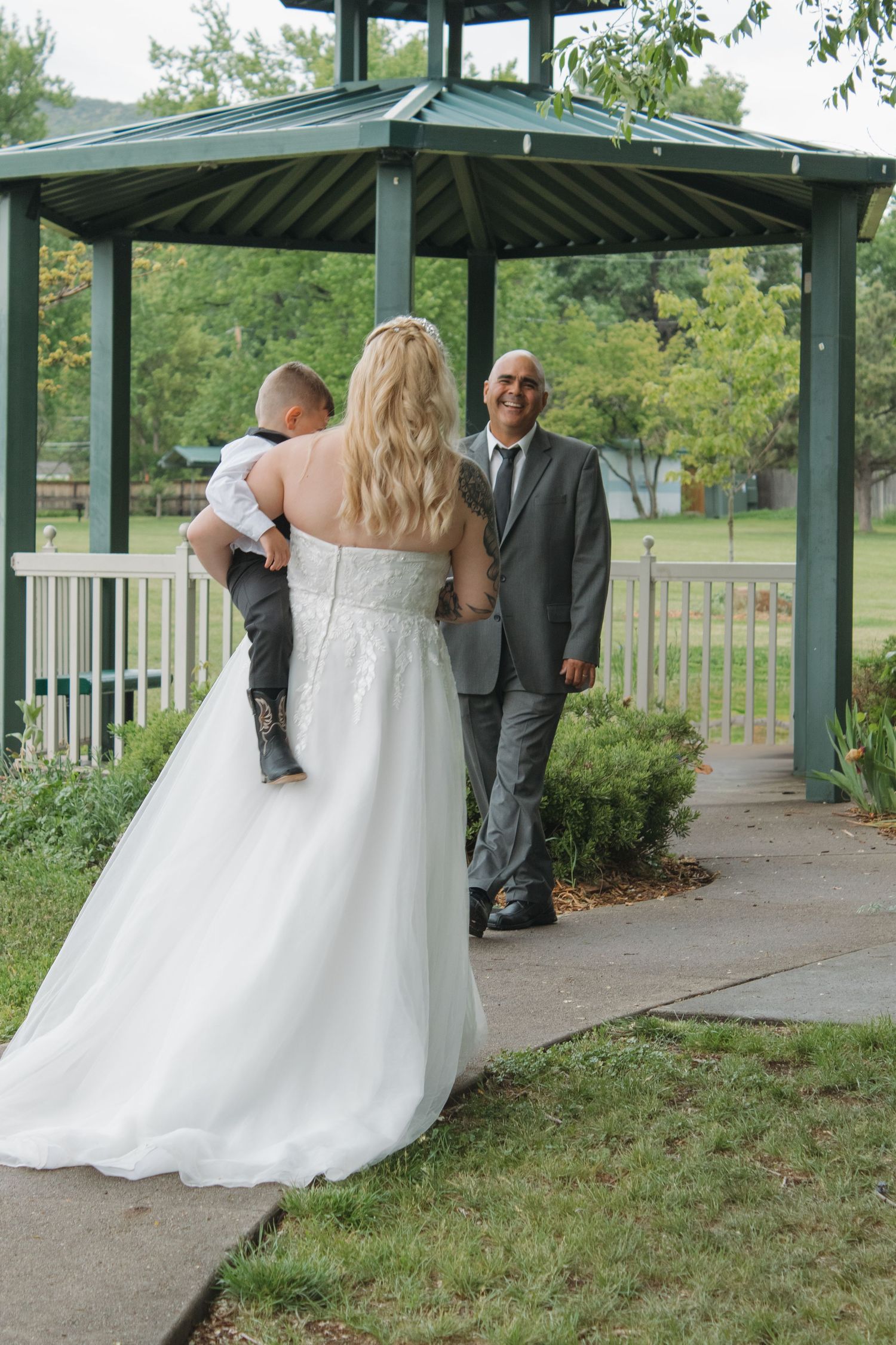 A bride in a flowing white gown walks under a green gazebo during an outdoor ceremony.