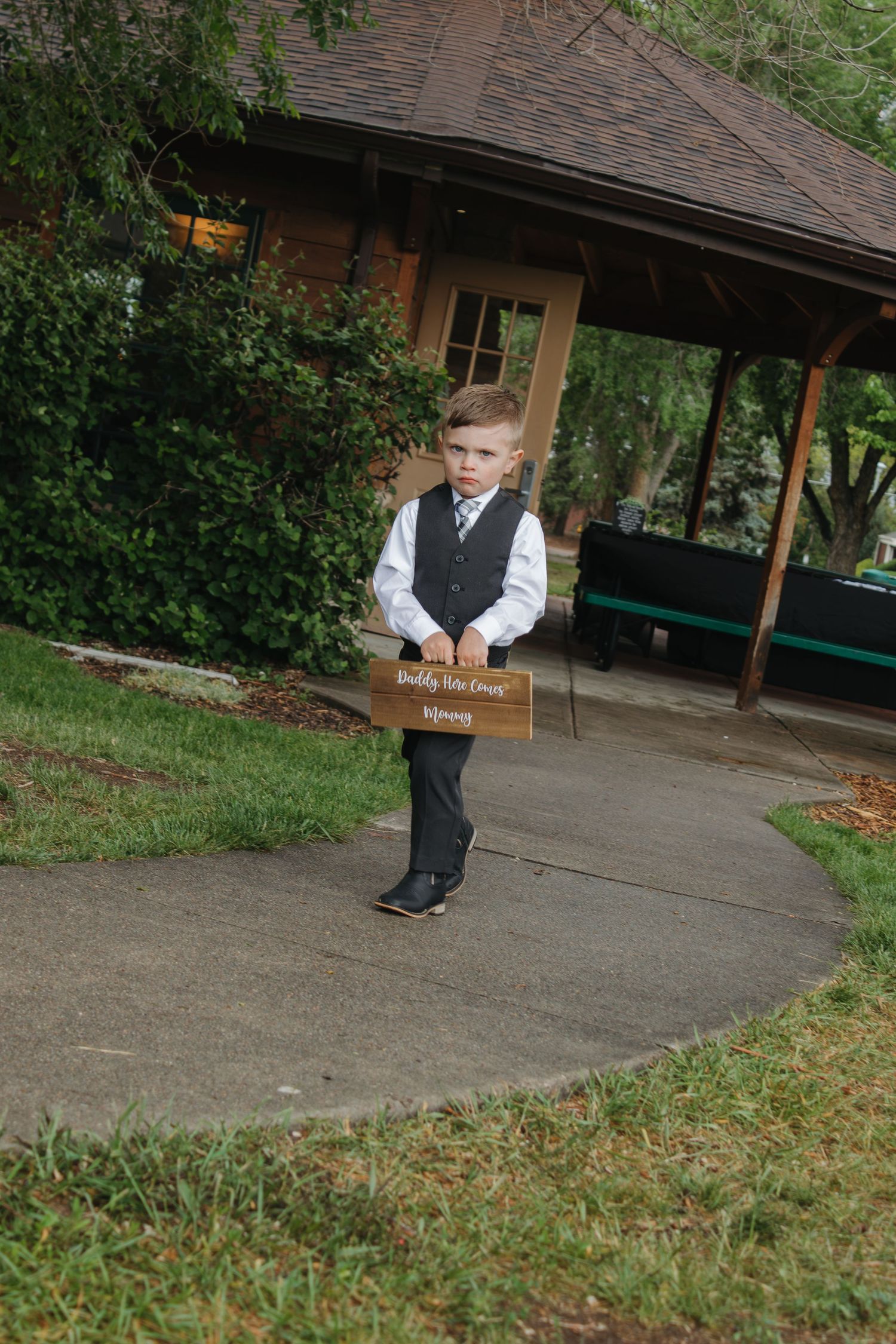 Young ring bearer in formal attire walking along covered outdoor pathway.