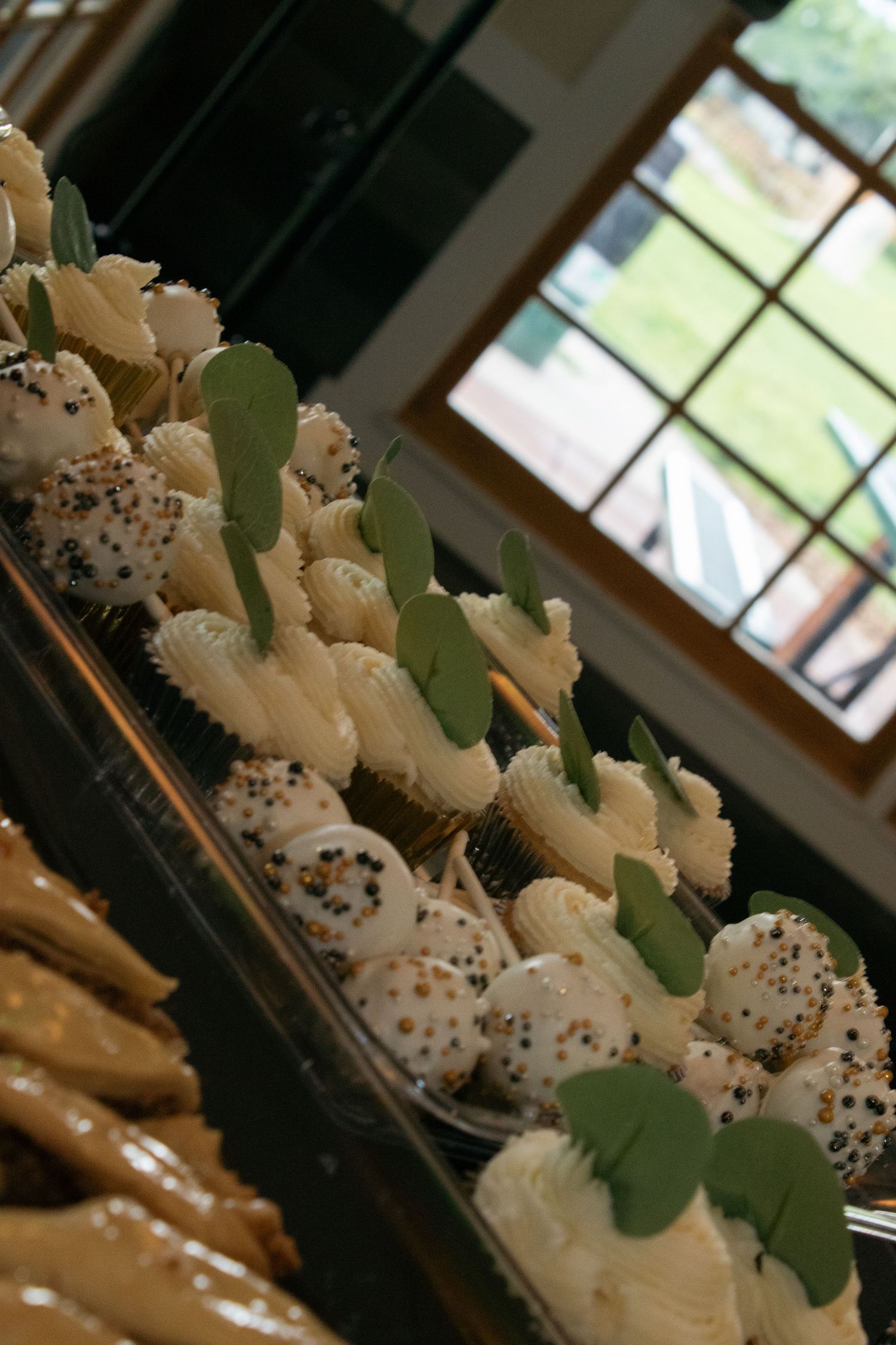 Elegant white frosted cupcakes decorated with sprinkles and green leaf accents displayed in rows.