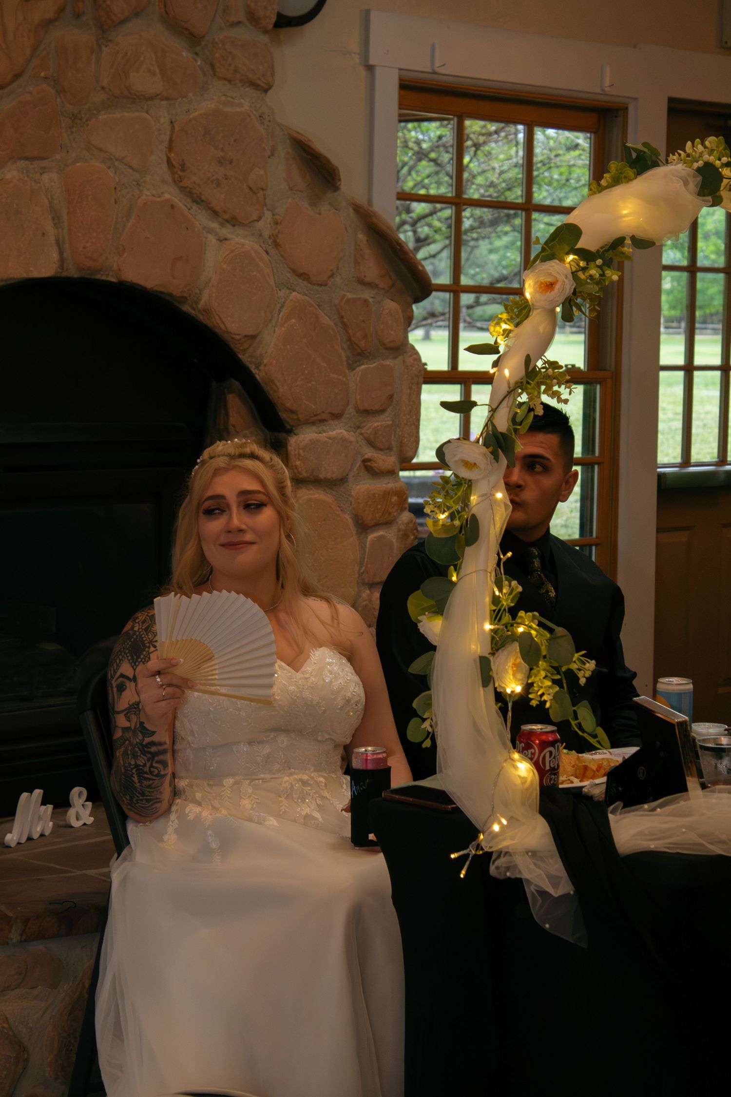 Wedding celebration by stone fireplace with decorative floral arch and paper fans.