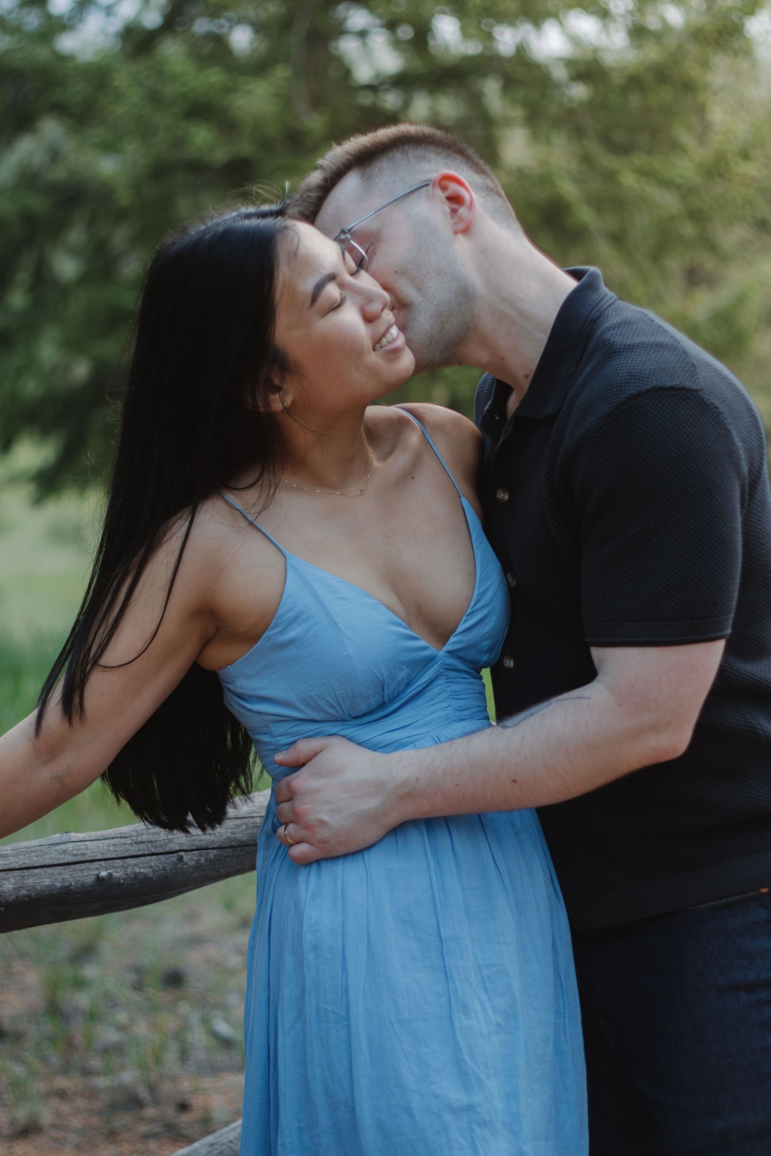 A loving couple shares a laugh while embracing near a wooden fence in nature.