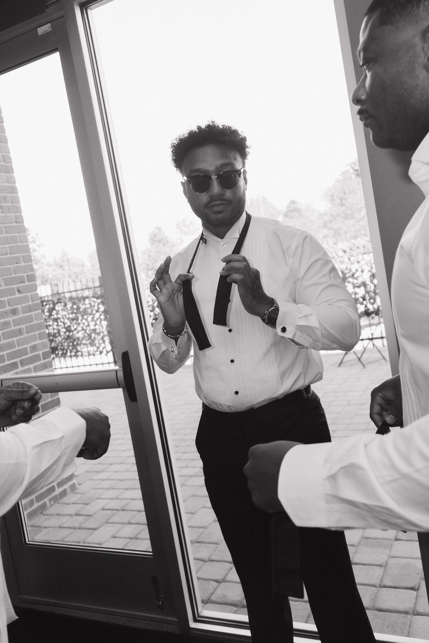Black and white photo of a groom in a tuxedo adjusting his tie near a glass door with groomsmen.
