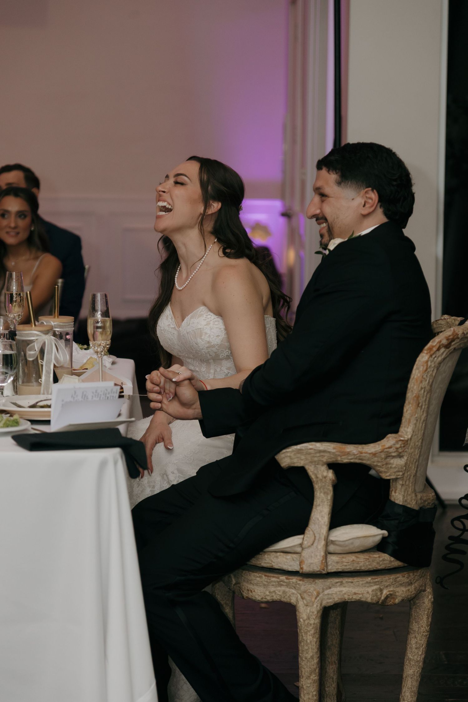 Bride and groom laughing joyfully while holding hands at their wedding reception sweetheart table.
