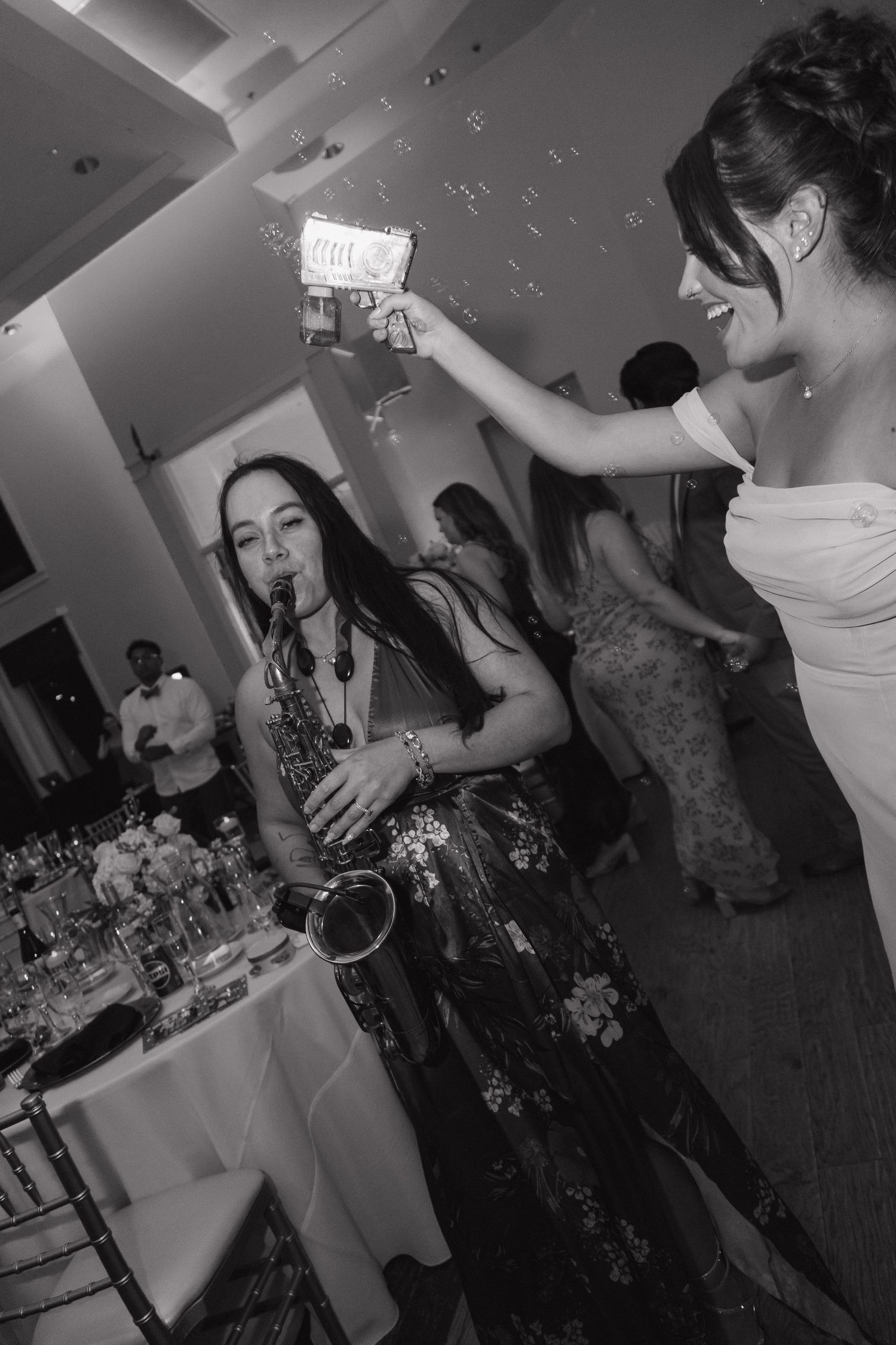 Black and white photo of a saxophonist performing at a wedding reception while a guest raises a drink.