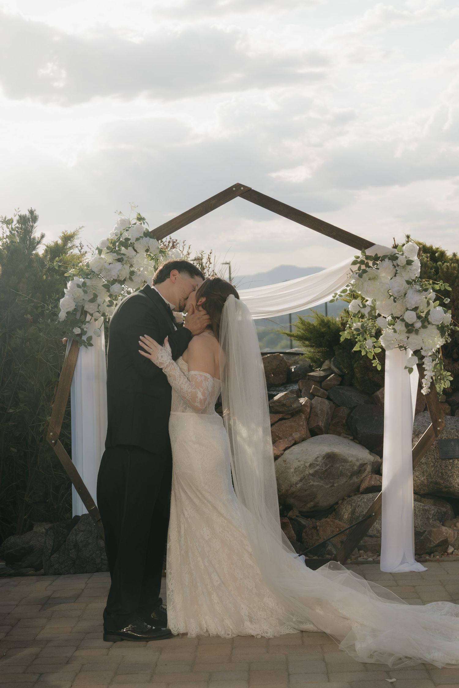 Bride and groom share first kiss under floral arch with mountain views.