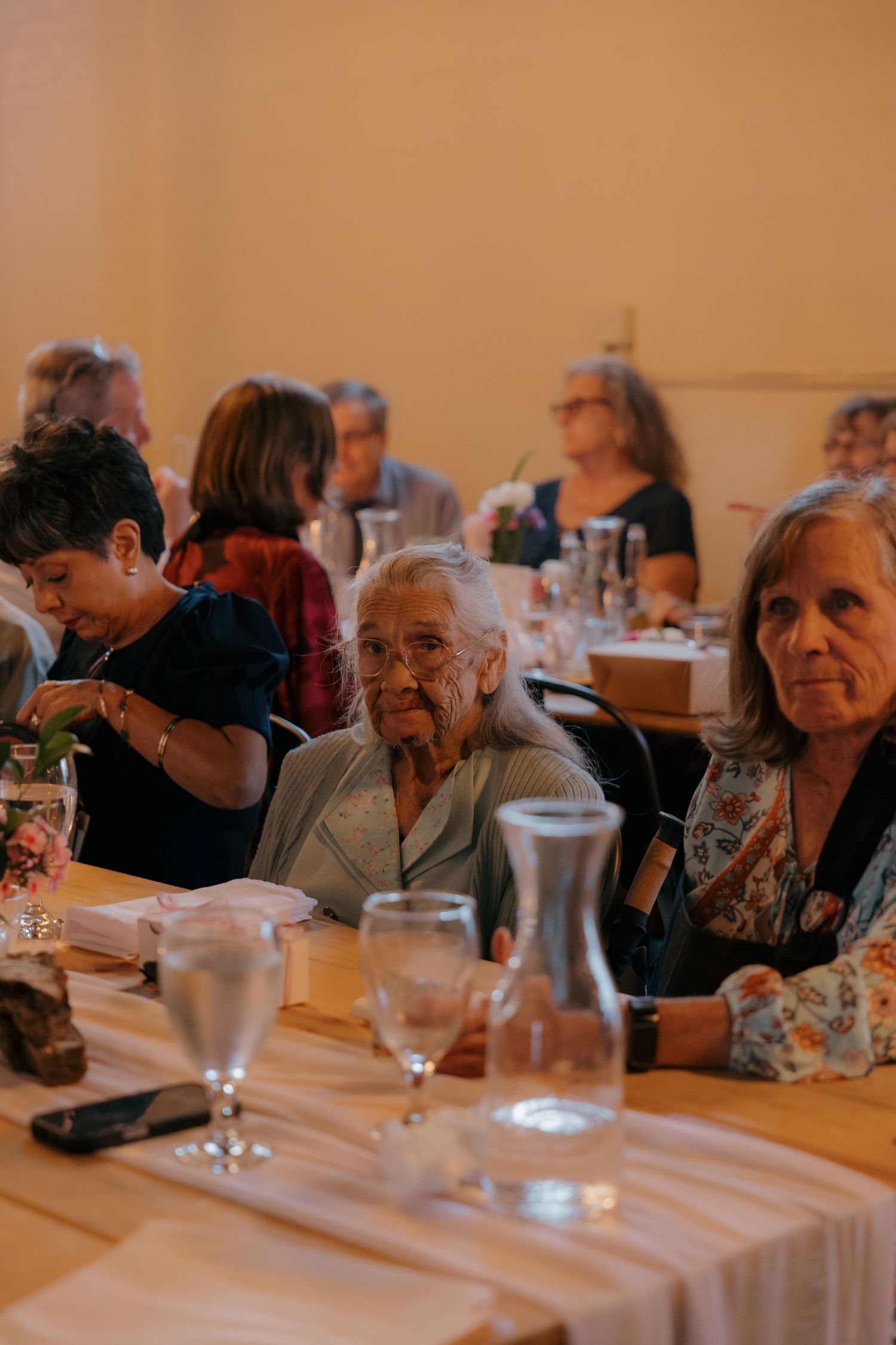 Elderly woman seated at wedding reception dinner table surrounded by other guests with water glasses nearby.