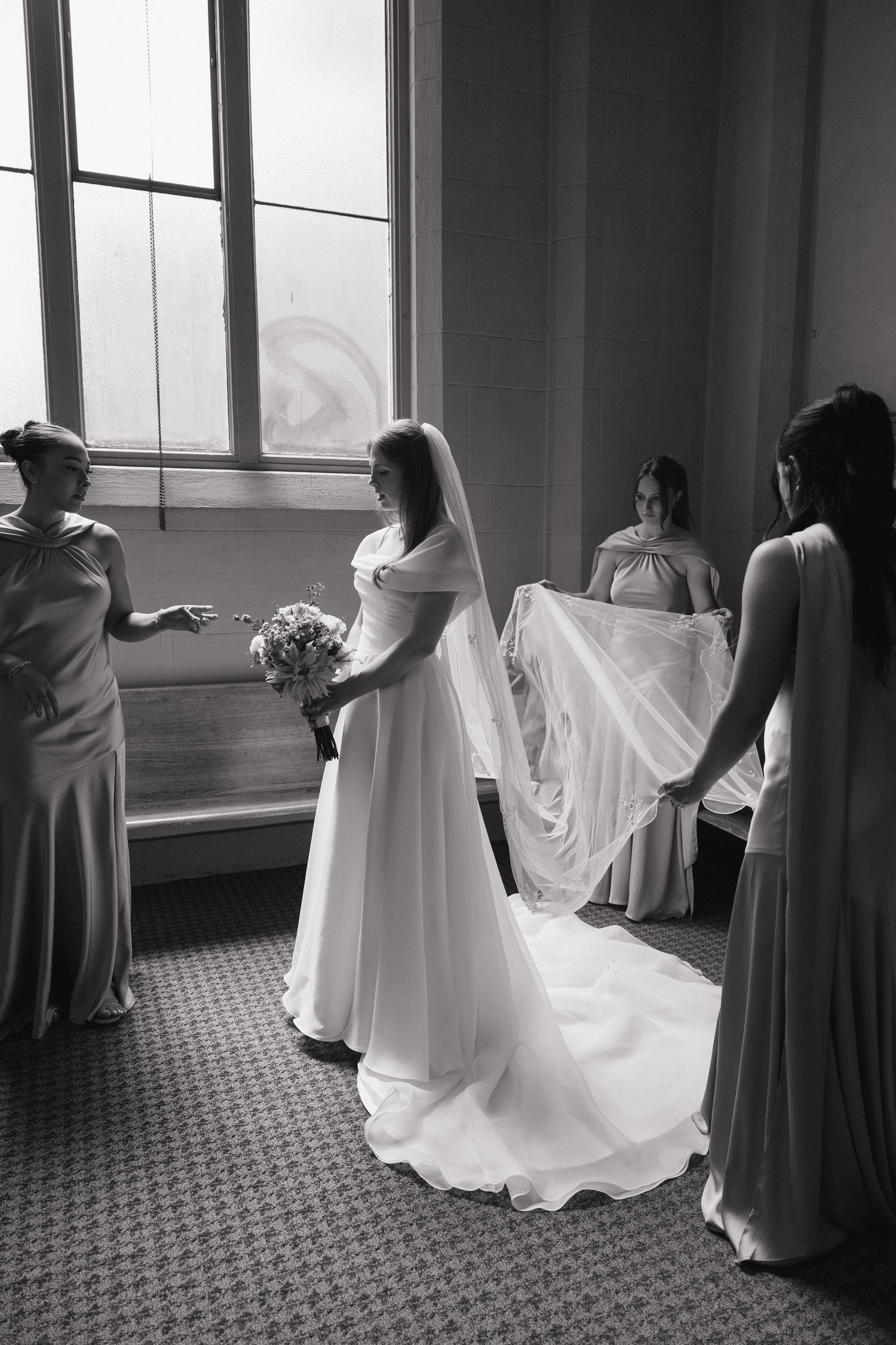 Bride in white gown and veil holds bouquet while bridesmaids adjust her train in black and white photo.