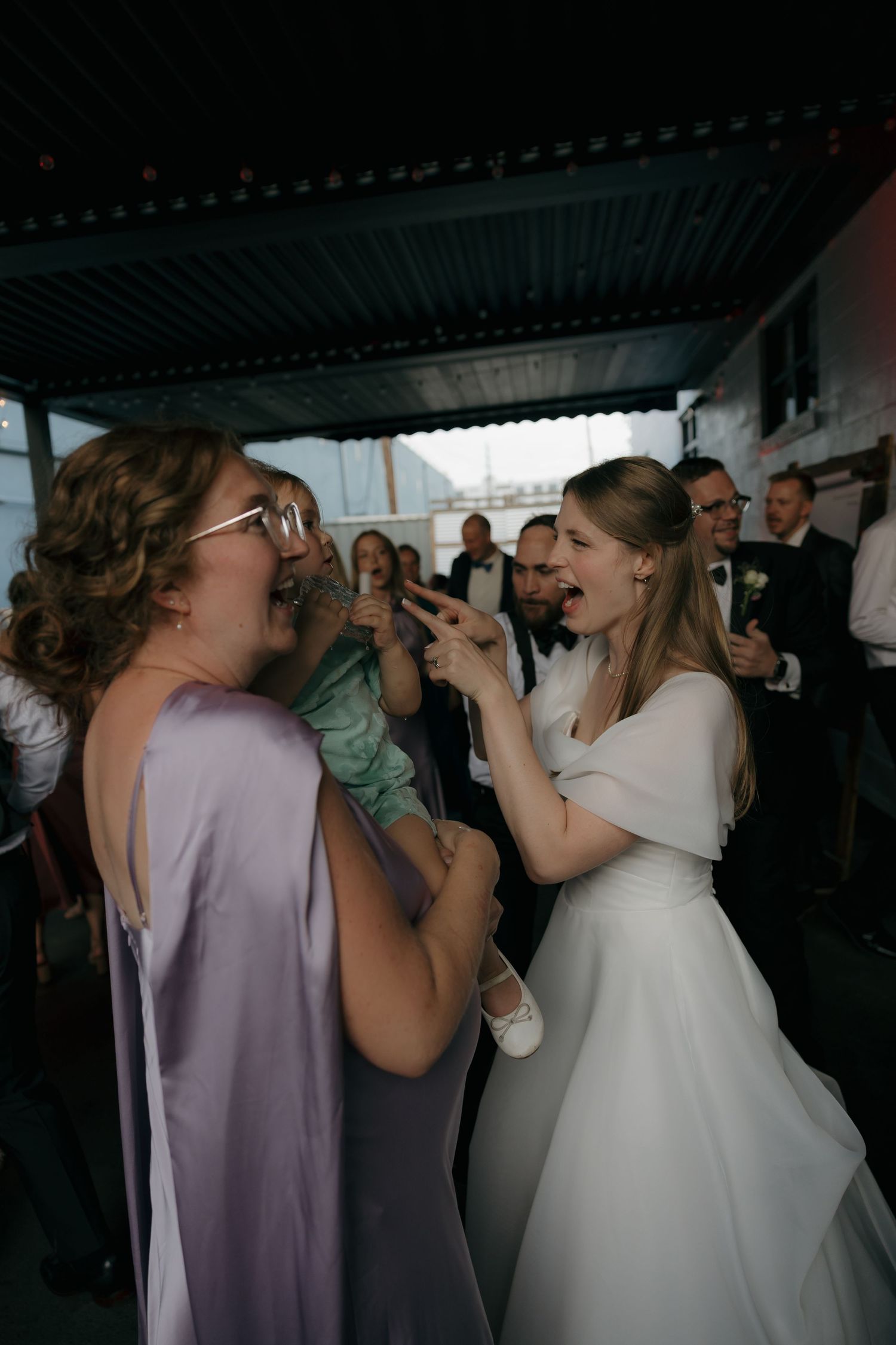 Bride in white gown laughs and dances with bridesmaid in lavender dress at wedding reception.