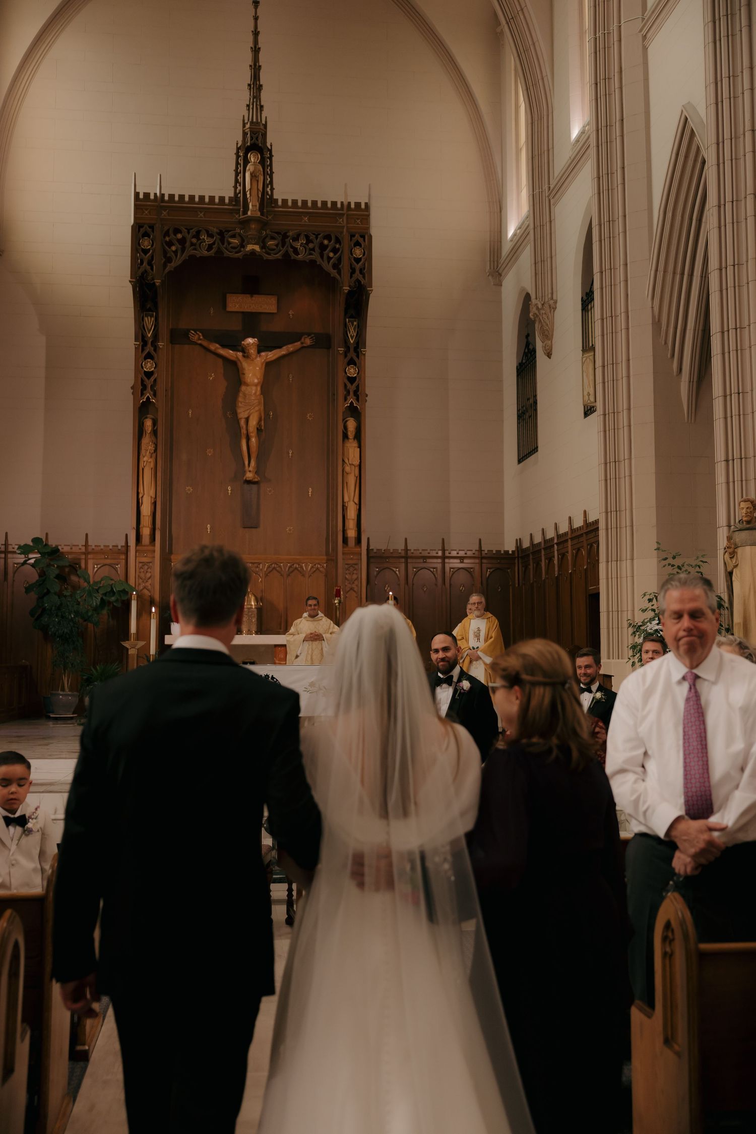 Bride and groom walking toward ornate crucifix altar in church accompanied by family during wedding ceremony.