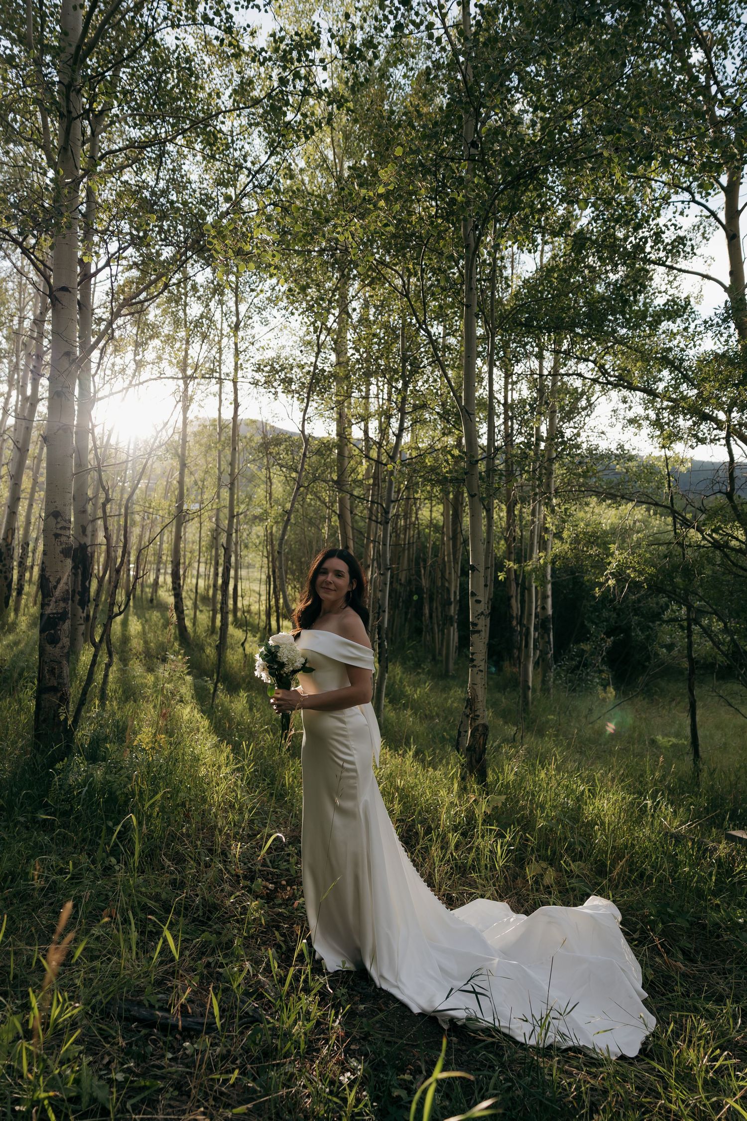 Bride in white off-shoulder gown with train stands in golden sunlit aspen forest holding bouquet.