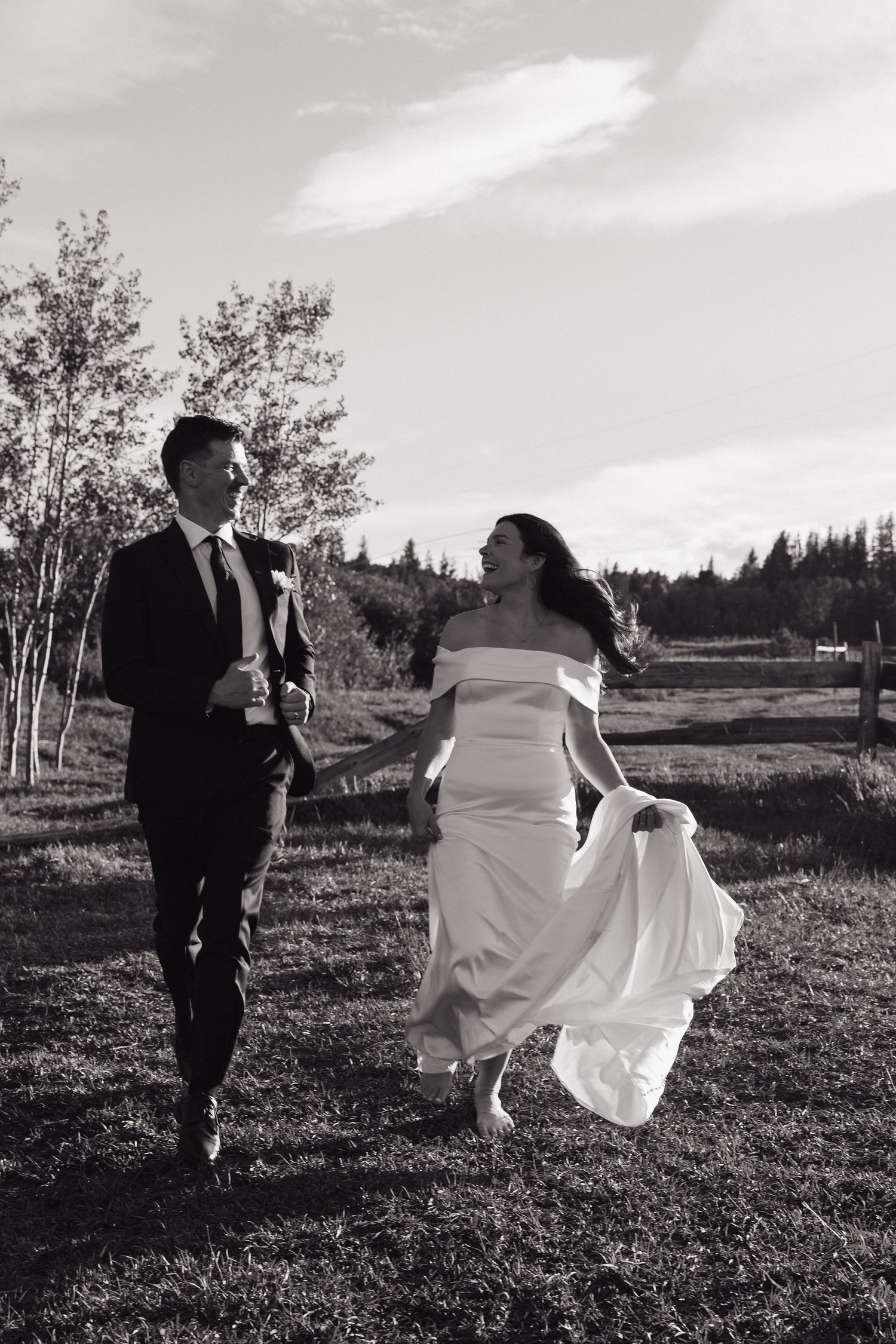 Black and white photo of bride and groom holding hands running through field at outdoor mountain wedding.