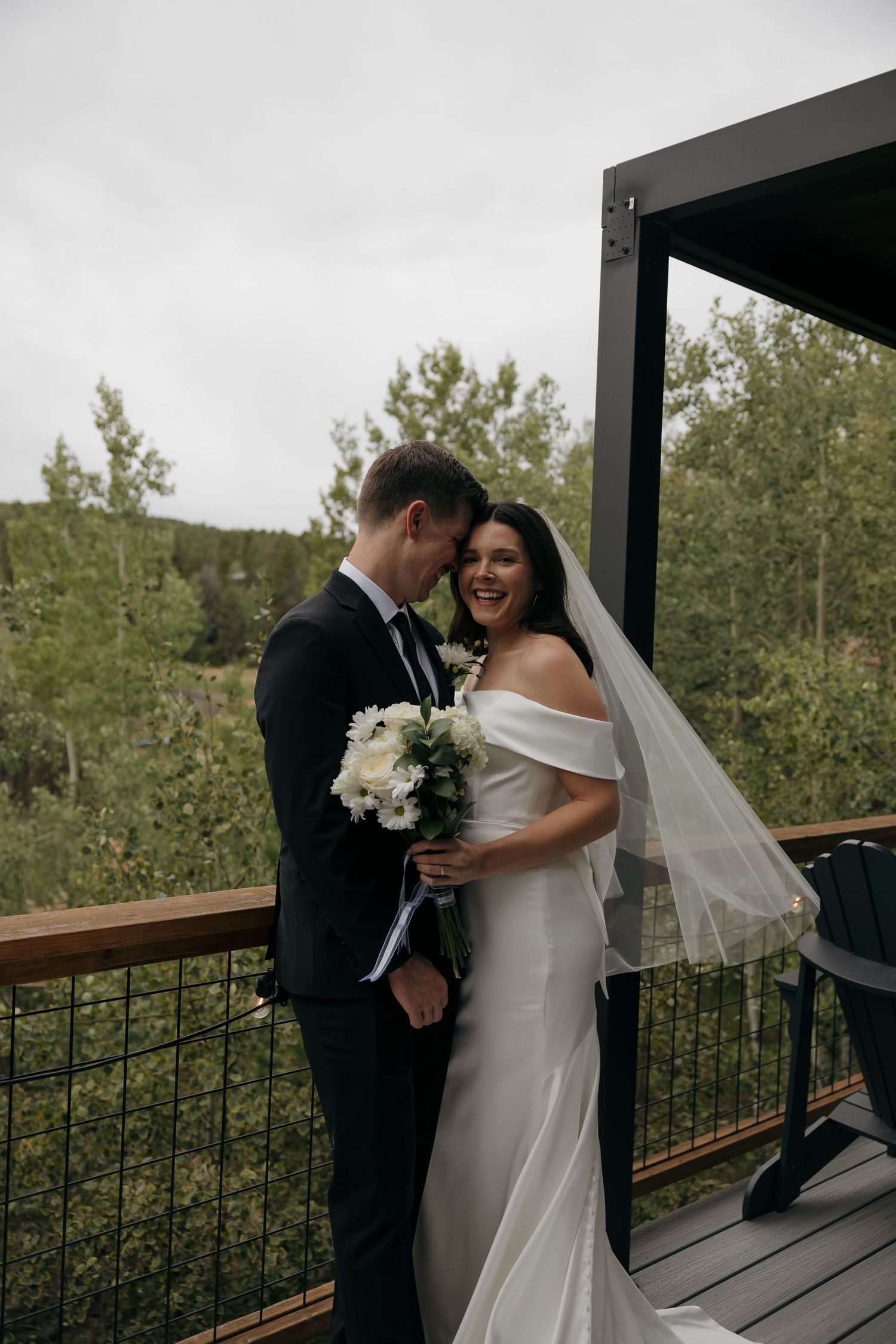 Laughing bride in off-shoulder gown with veil embraces groom on wooden deck surrounded by trees.