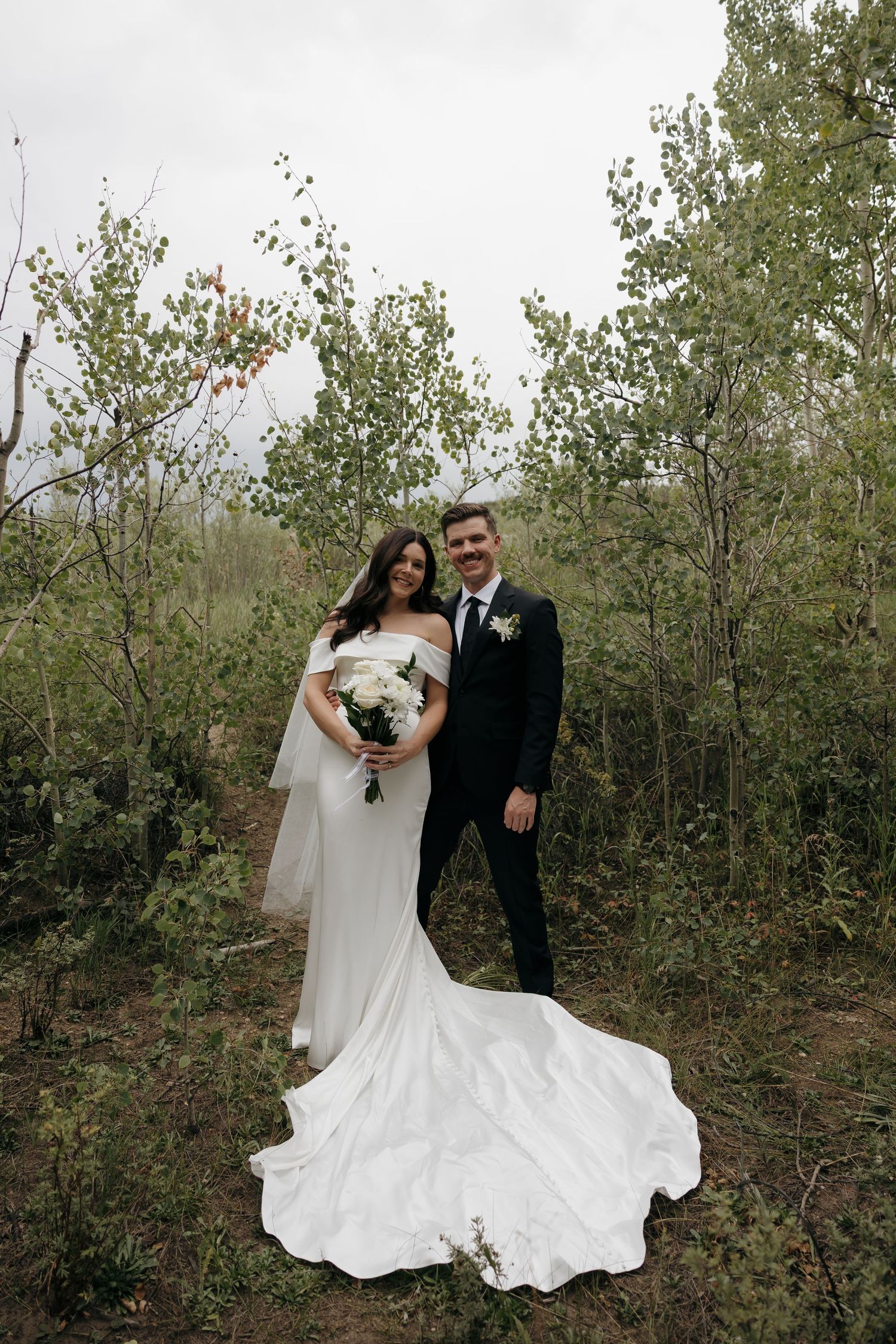 Bride and groom pose together in a forest setting, she in a white off-shoulder gown holding a bouquet, he in a black suit.