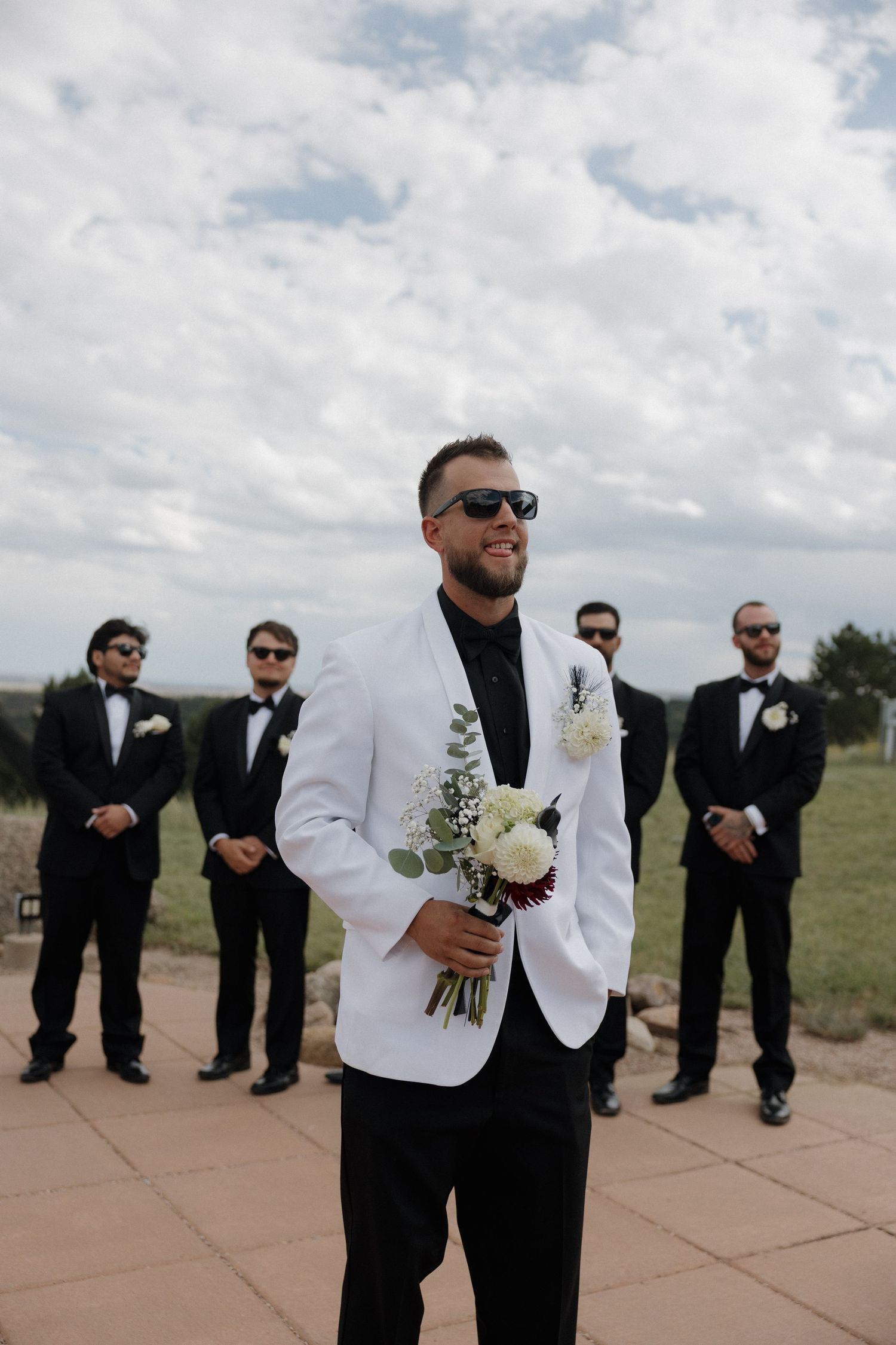 Groom in white tuxedo jacket holds bouquet with groomsmen in black suits behind him outdoors.