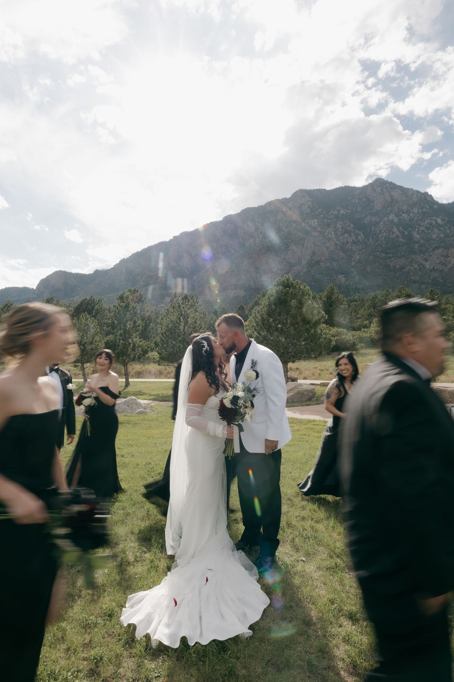Bride and groom share a kiss outdoors with mountain backdrop, bridesmaids in black dresses surrounding them.