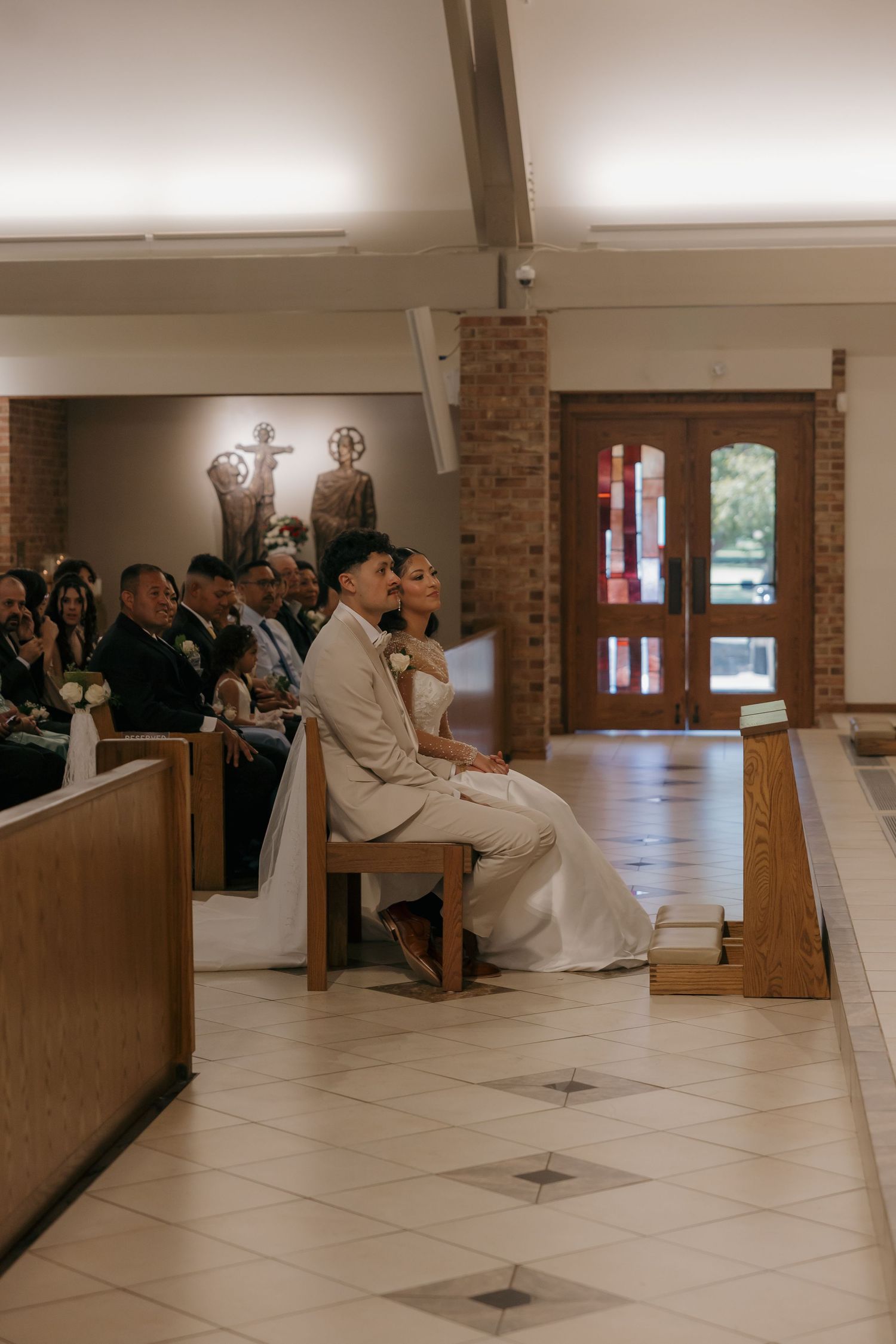 Bride and groom seated at the altar during a church wedding ceremony, with guests in pews and religious artwork on the walls.