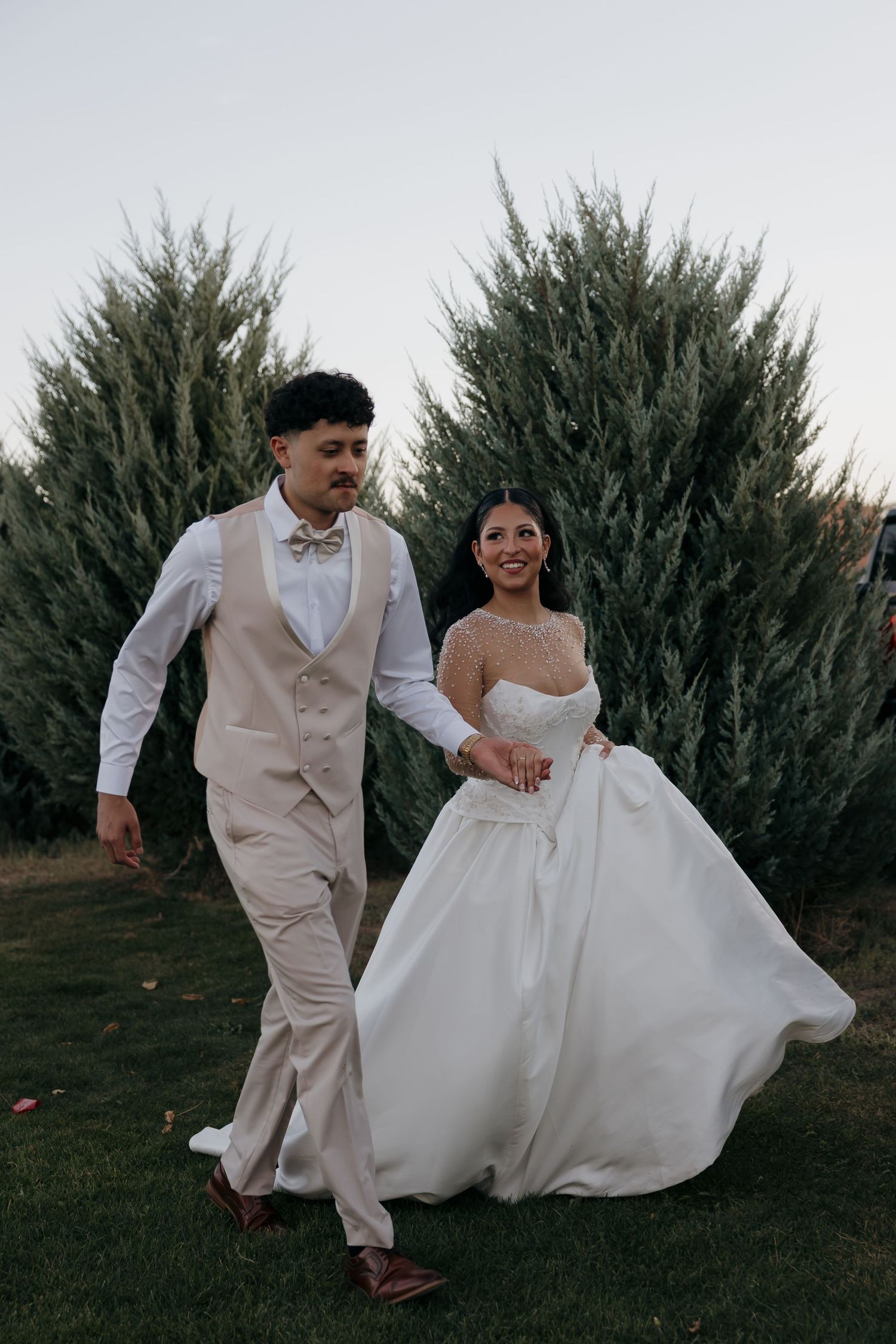 Bride in lace-sleeved white gown and groom in tan vest and bow tie walk hand in hand before evergreen trees at dusk.