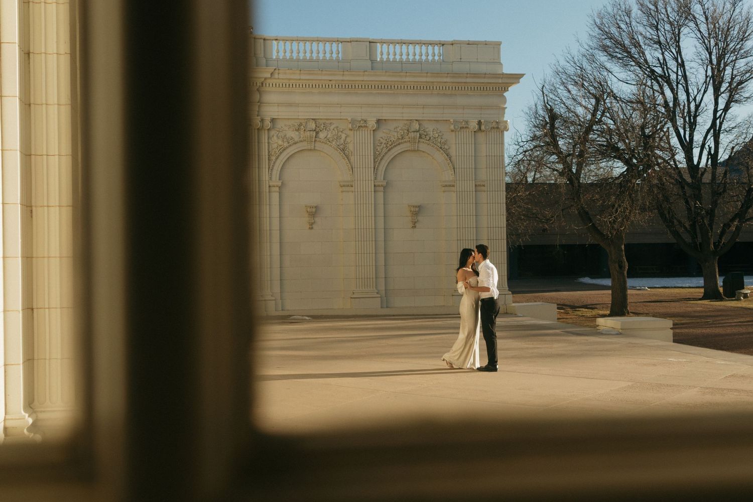 A couple embraces in front of classical architecture with arches and columns on a bright day.