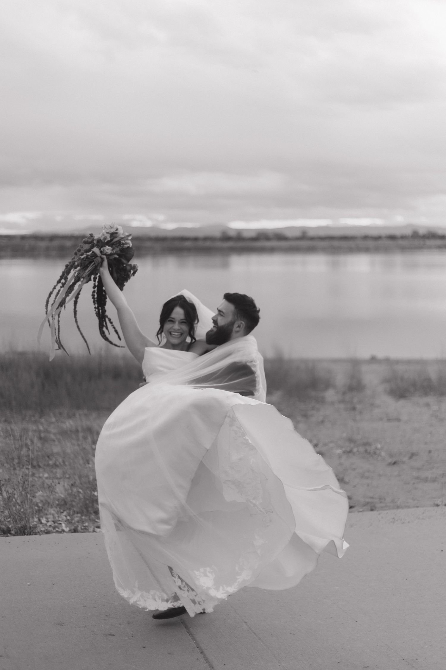 Ethereal wedding photos by a lake with bride's dress flowing dramatically in the wind.