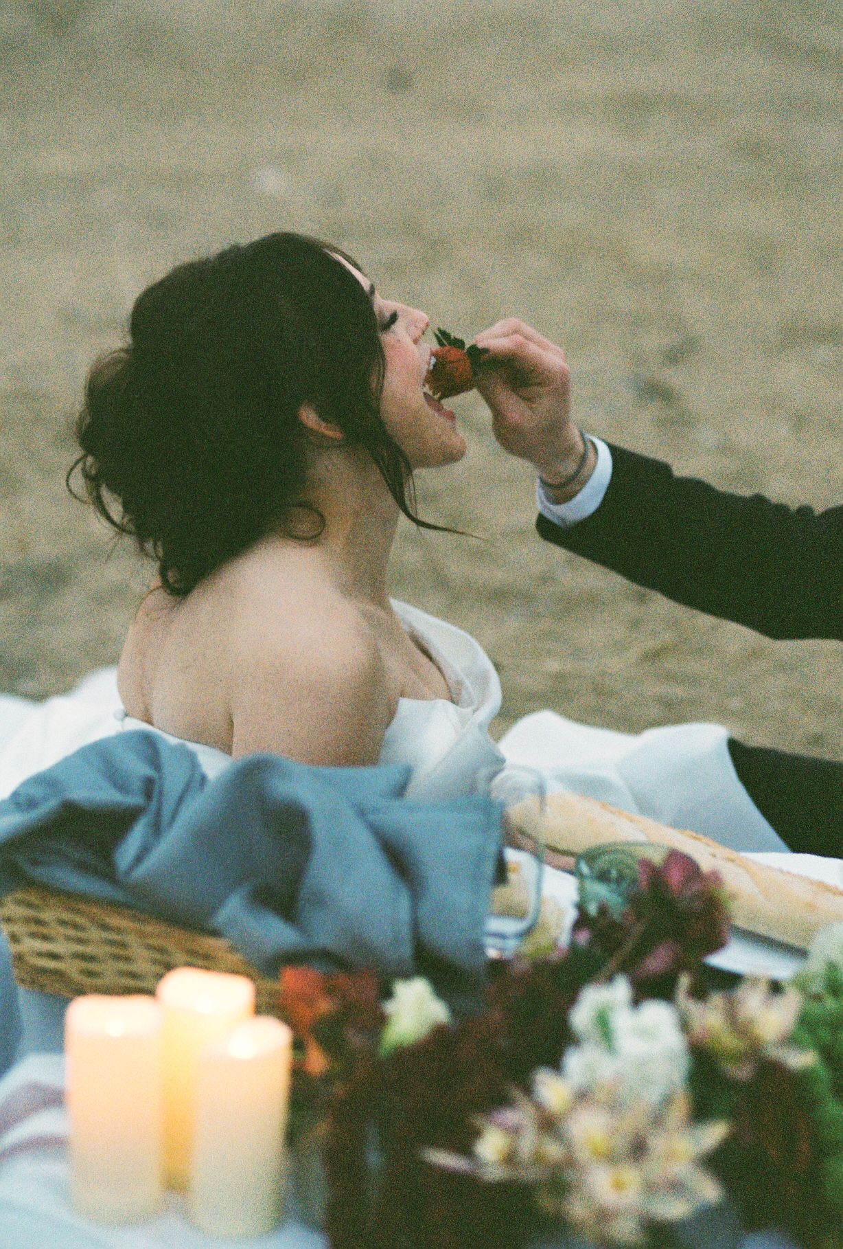 Couple shares romantic moment on beach blanket with candles and flowers at sunset.
