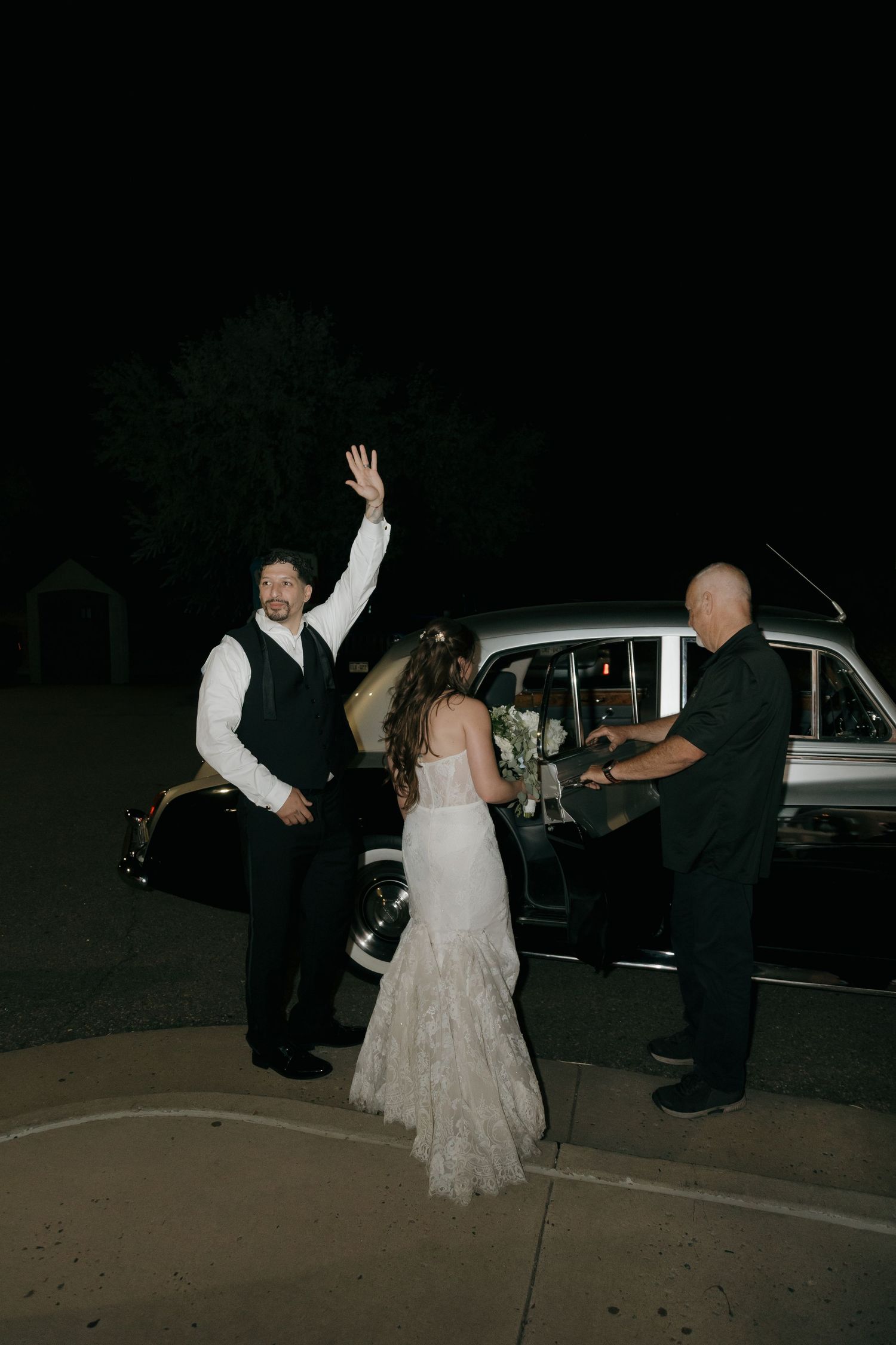 Groom waves beside classic vintage car as bride and driver stand nearby at nighttime wedding exit.