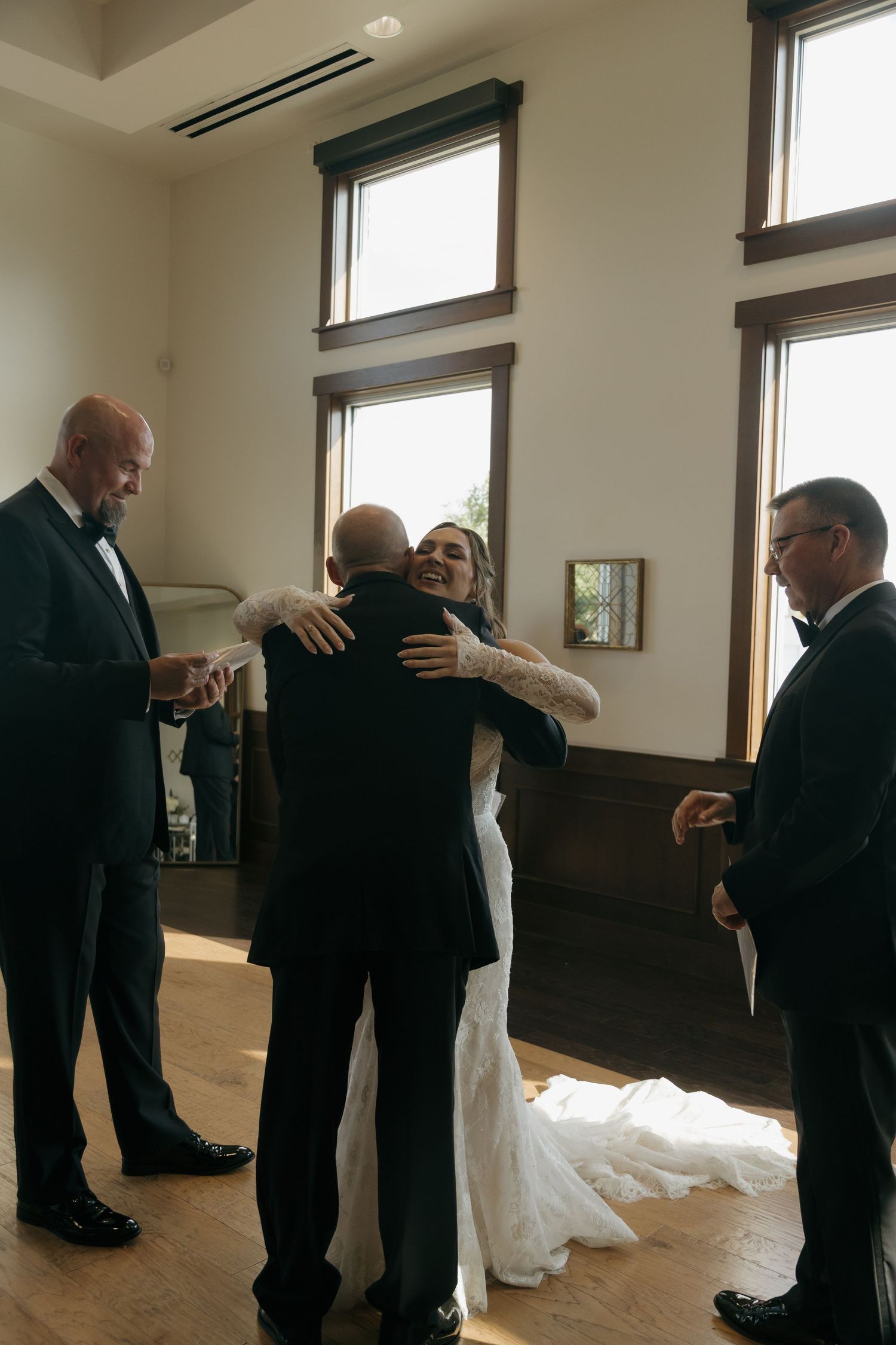 Emotional bride hugging guest in elegant wedding venue with wood floors and tall windows.