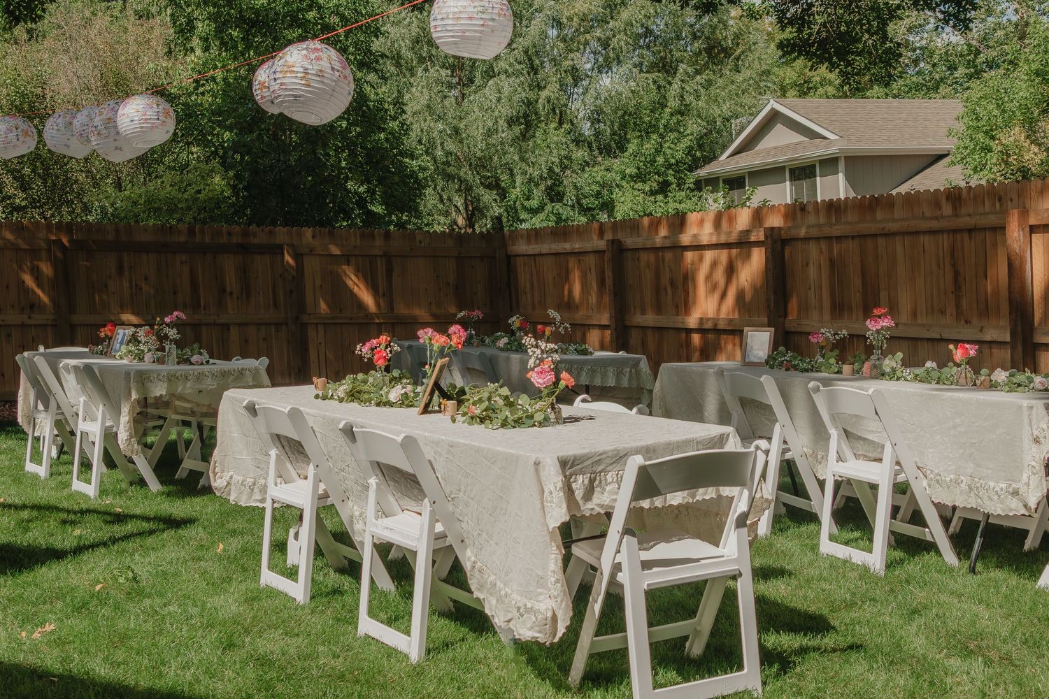 An elegant outdoor garden wedding reception setup with white tables and chairs decorated with floral arrangements and lanterns.