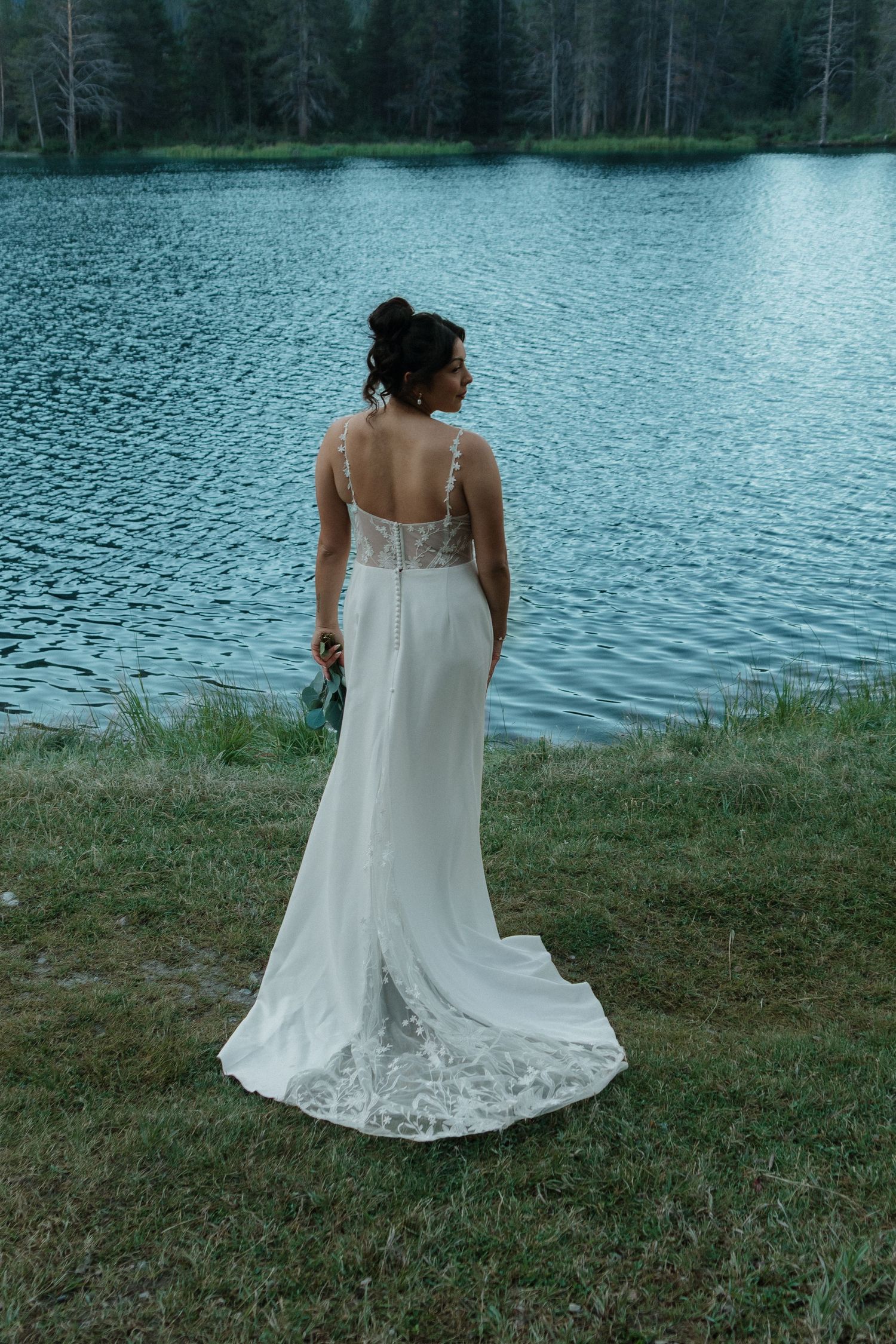 A person in a white wedding dress stands near a serene lake surrounded by mountains.