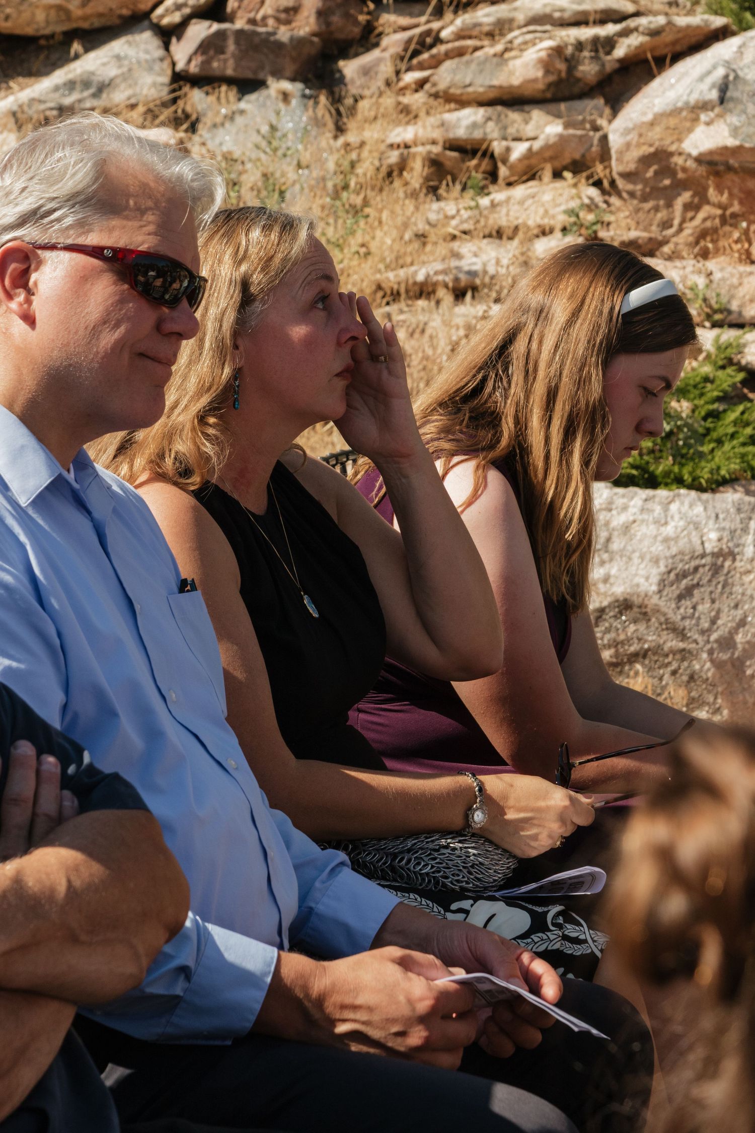 Wedding guests seated on stone benches watching ceremony in bright sunlight.