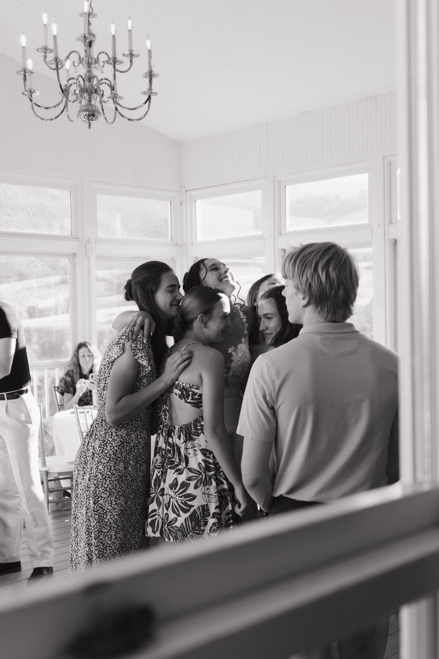 Black and white photo of wedding reception guests mingling under chandelier.