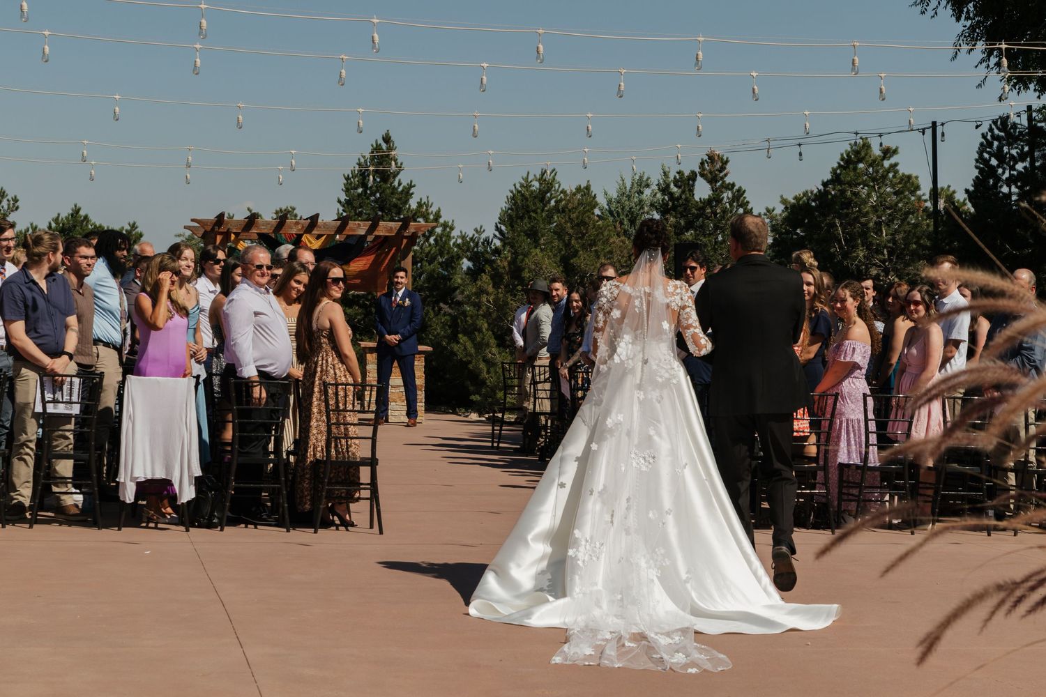 Wedding ceremony guests seated outdoors under string lights on a sunny day.