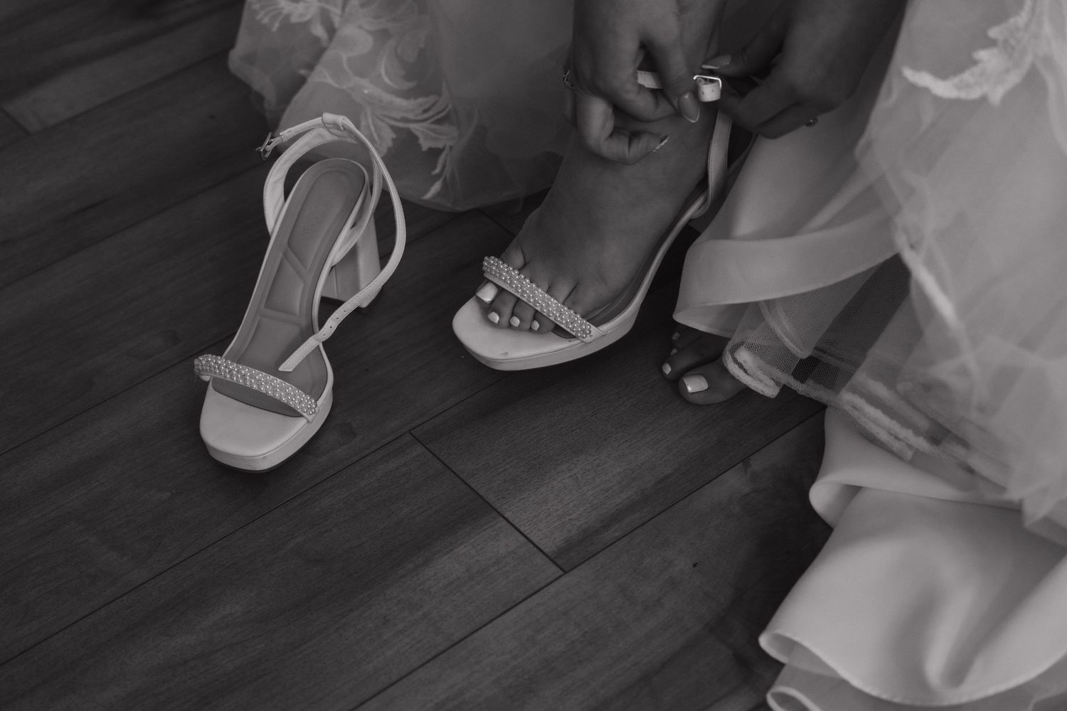 Black and white close-up of bride putting on embellished strappy wedding heels on wooden floor.
