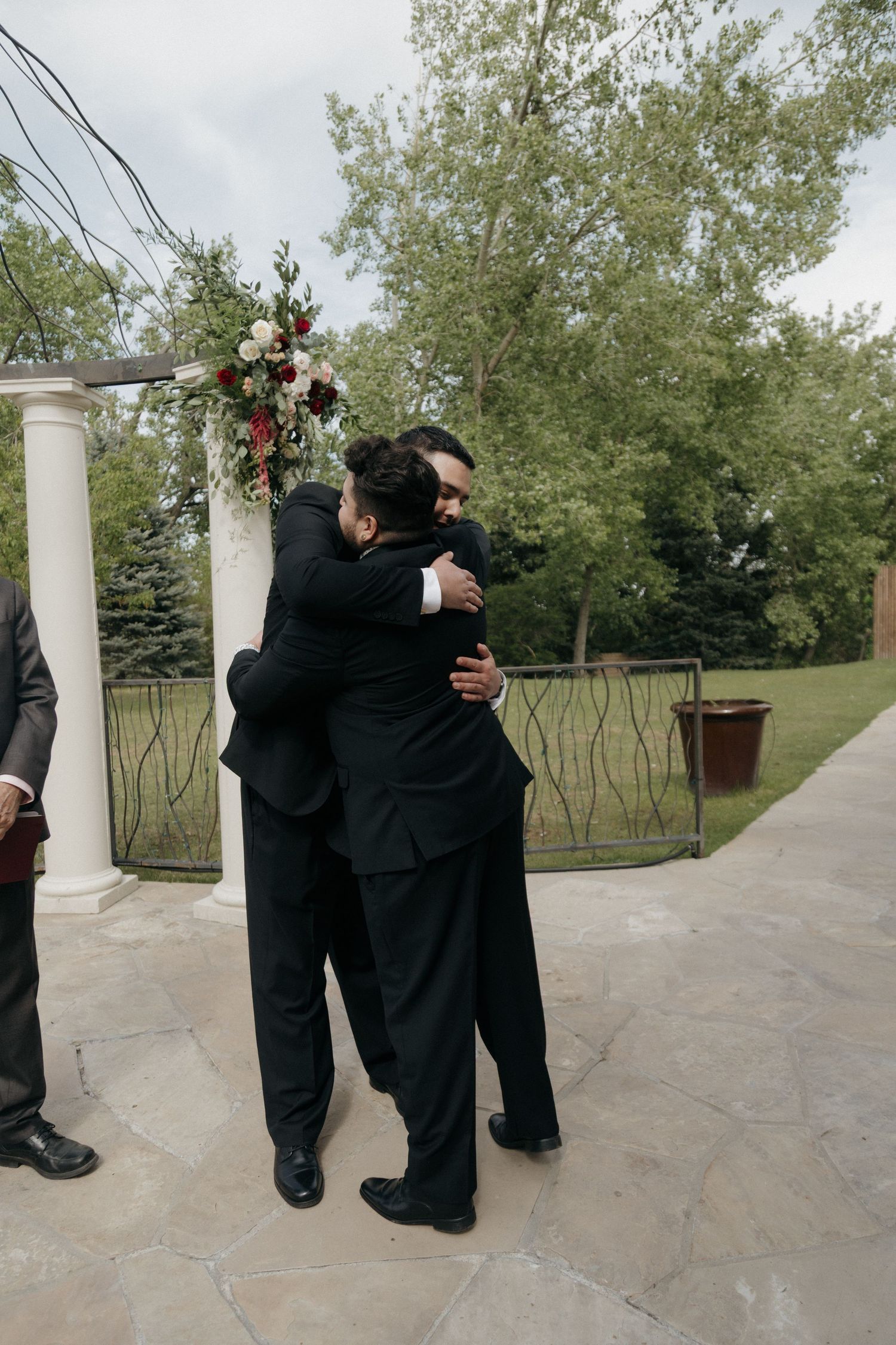 Two groomsmen in black tuxedos share an emotional hug near floral-decorated wedding ceremony arch outdoors.